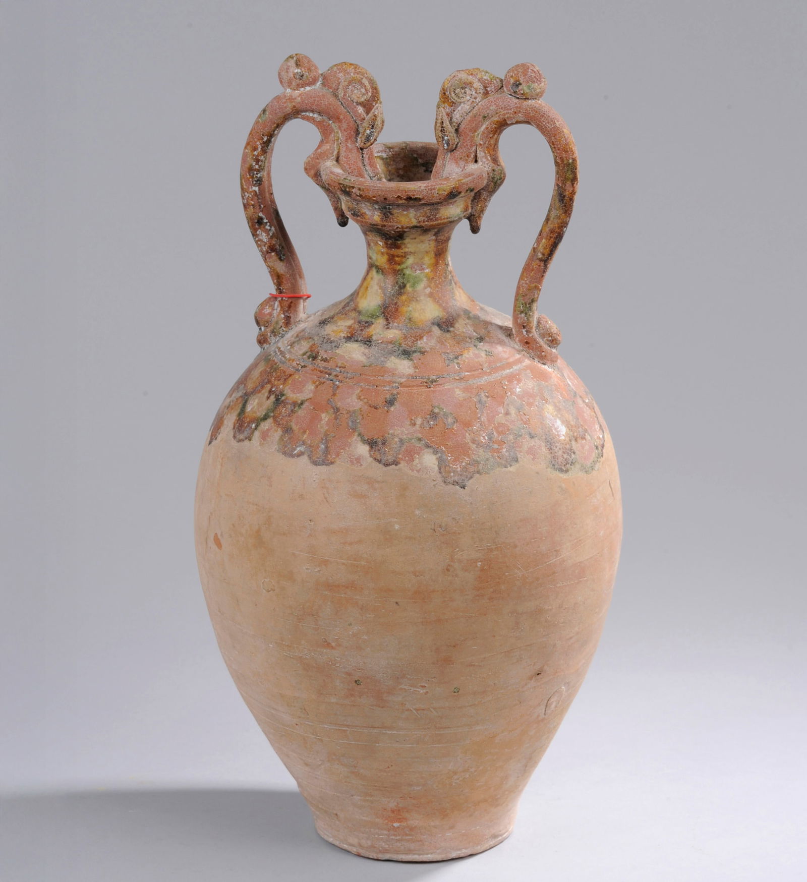 Sancai glazed amphora covered with a tricolor glaze. China. Tang Dynasty.: An ovoid-shaped amphora on a narrow base and high neck, with two wide zoomorphic handles with dragon mouths, in partially glazed Sancai terracotta and covered with a tricolor glaze. China. Tang. 37 cm