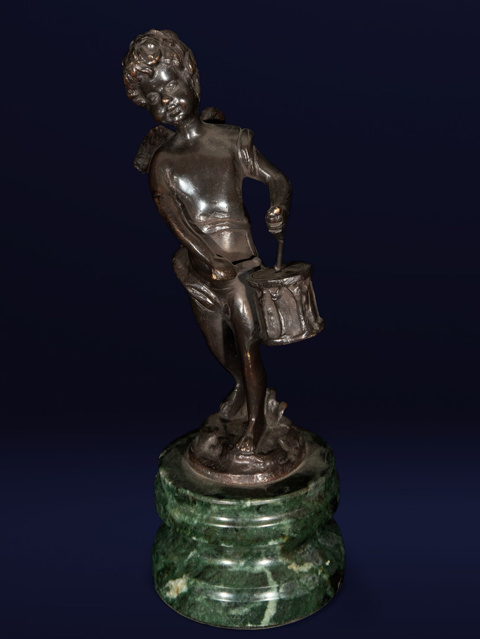 The Young Drummer Boy. European School. 19th Century.: Patinated bronze on a green marble base. Dimensions: 30 x 10 x 10 cm. The figure of a cherub is presented in a dynamic pose, holding a drum in one hand and a drumstick in the other, in the midst of mu