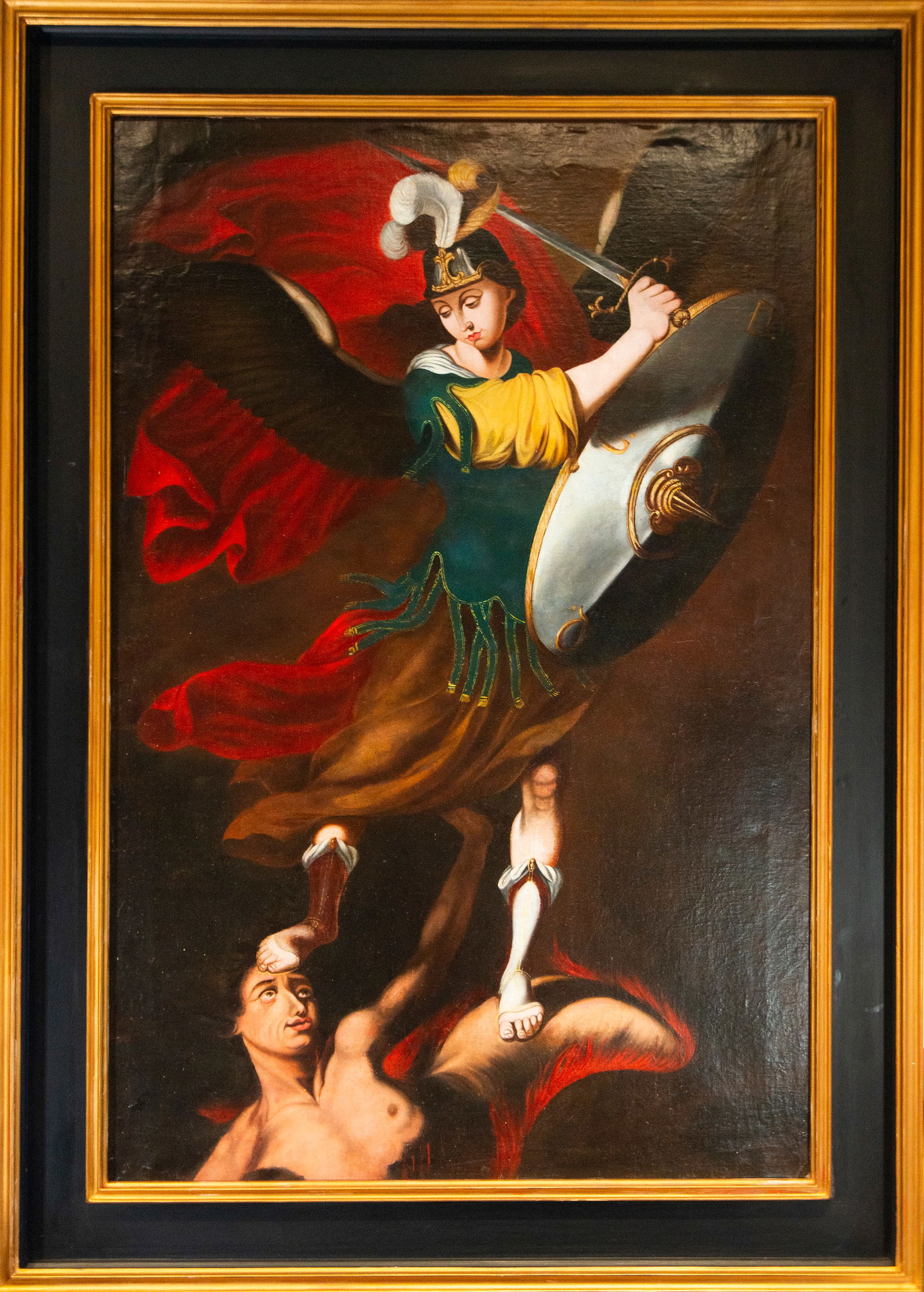 Archangel vanquished by the devil. Mexican Viceroyalty School. 18th Century.: Oil on canvas. Dimensions 150 × 100 cm. A winged angelic figure, in armor, with a feathered helmet and a large metal shield, rises triumphantly, subduing the defeated demon with its foot. The angel r