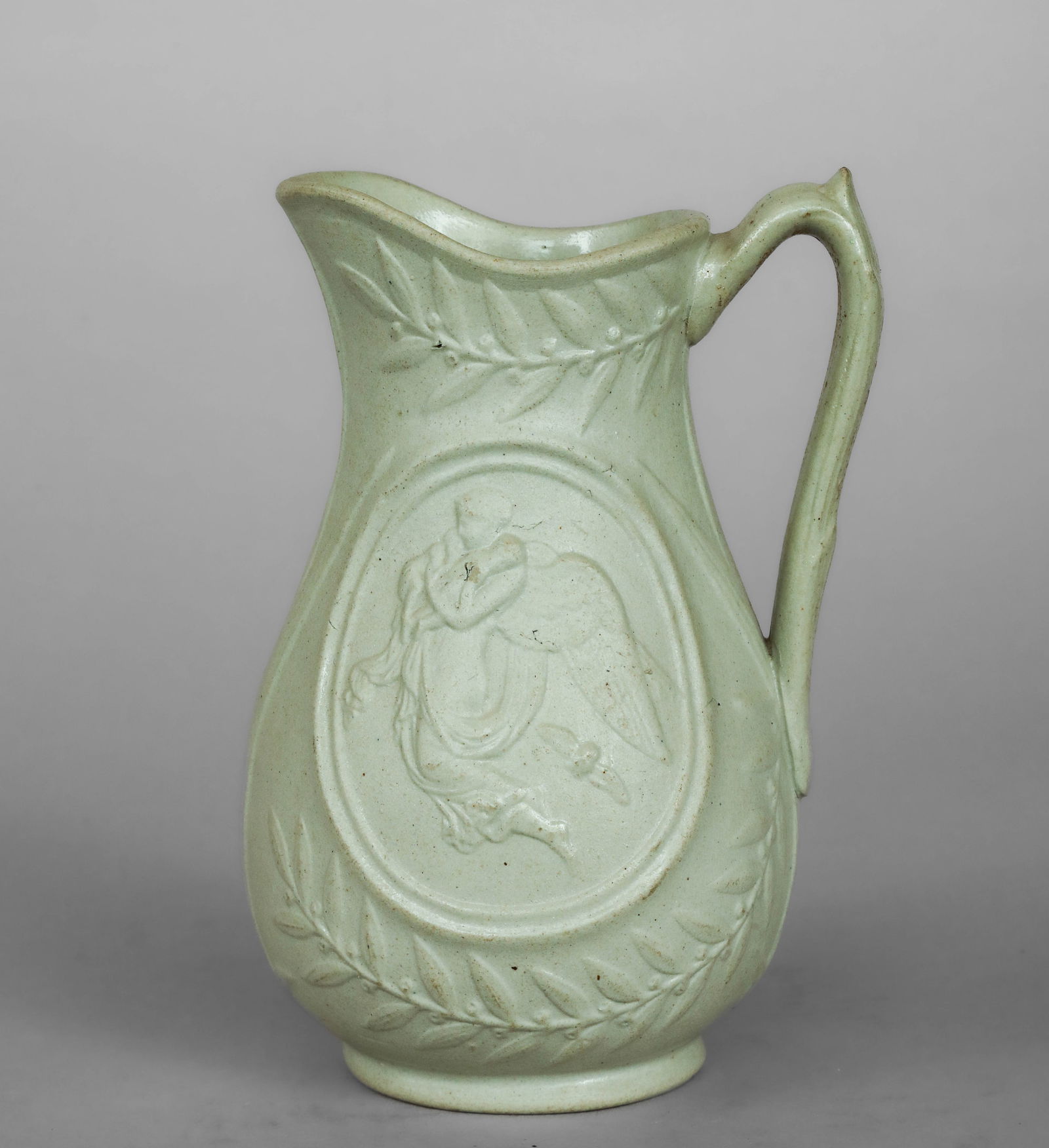 Pitcher with Angel and Olive Branches, James Dudson, England, 19th Century: Green monochrome glazed ceramic bisque. Measures 16 cm. A jug with a rounded body and side handle, decorated with a central angel motif set in an oval, surrounded by olive branches that follow the con