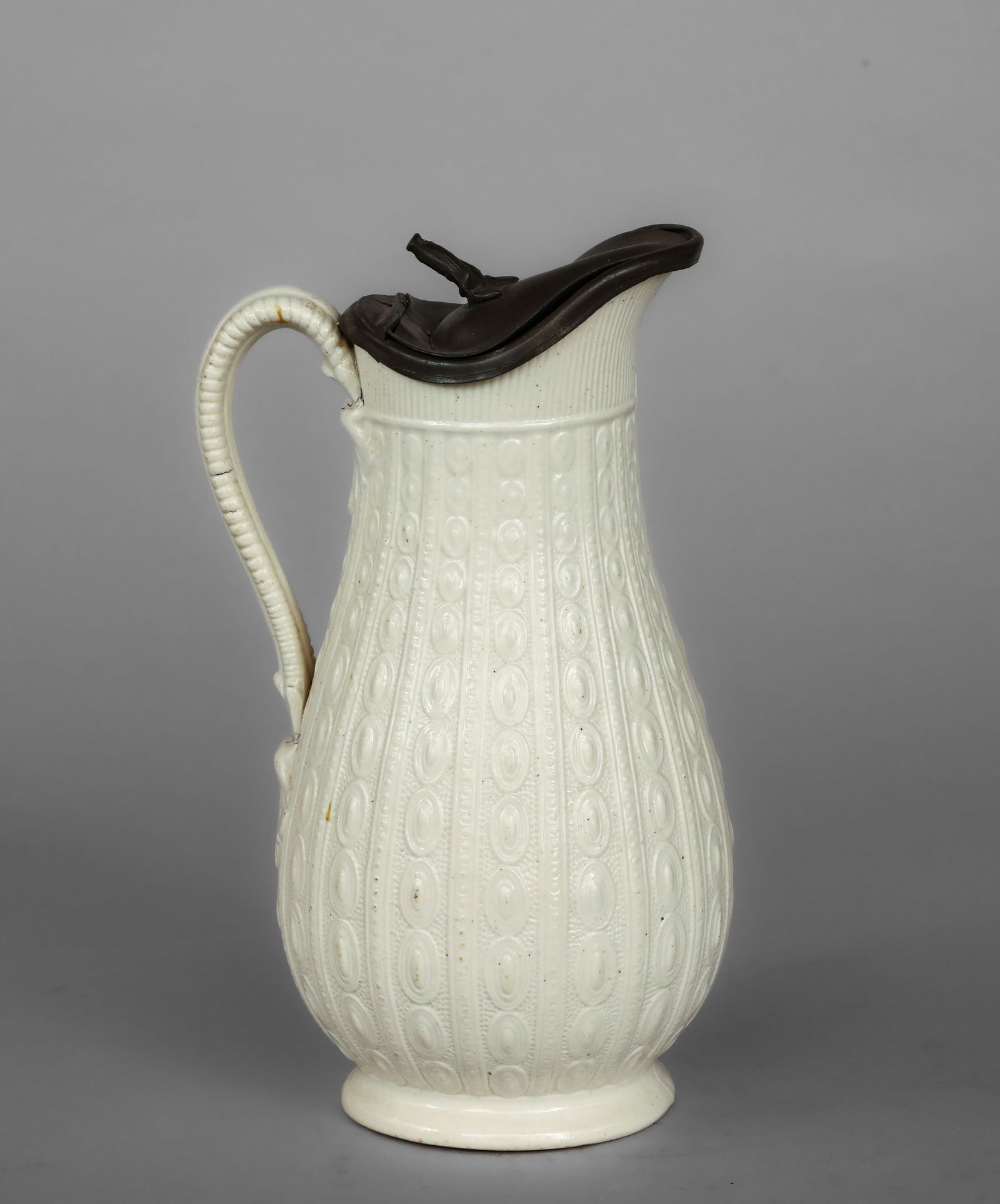 Jug with friezes of oval medallions and pewter lid, James Dudson, England, 19th century: White monochrome glazed ceramic bisque with a tin lid. Measures 22.5 cm in height. The slender jug ??features a side handle and is decorated with horizontal friezes of oval medallions in relief that r