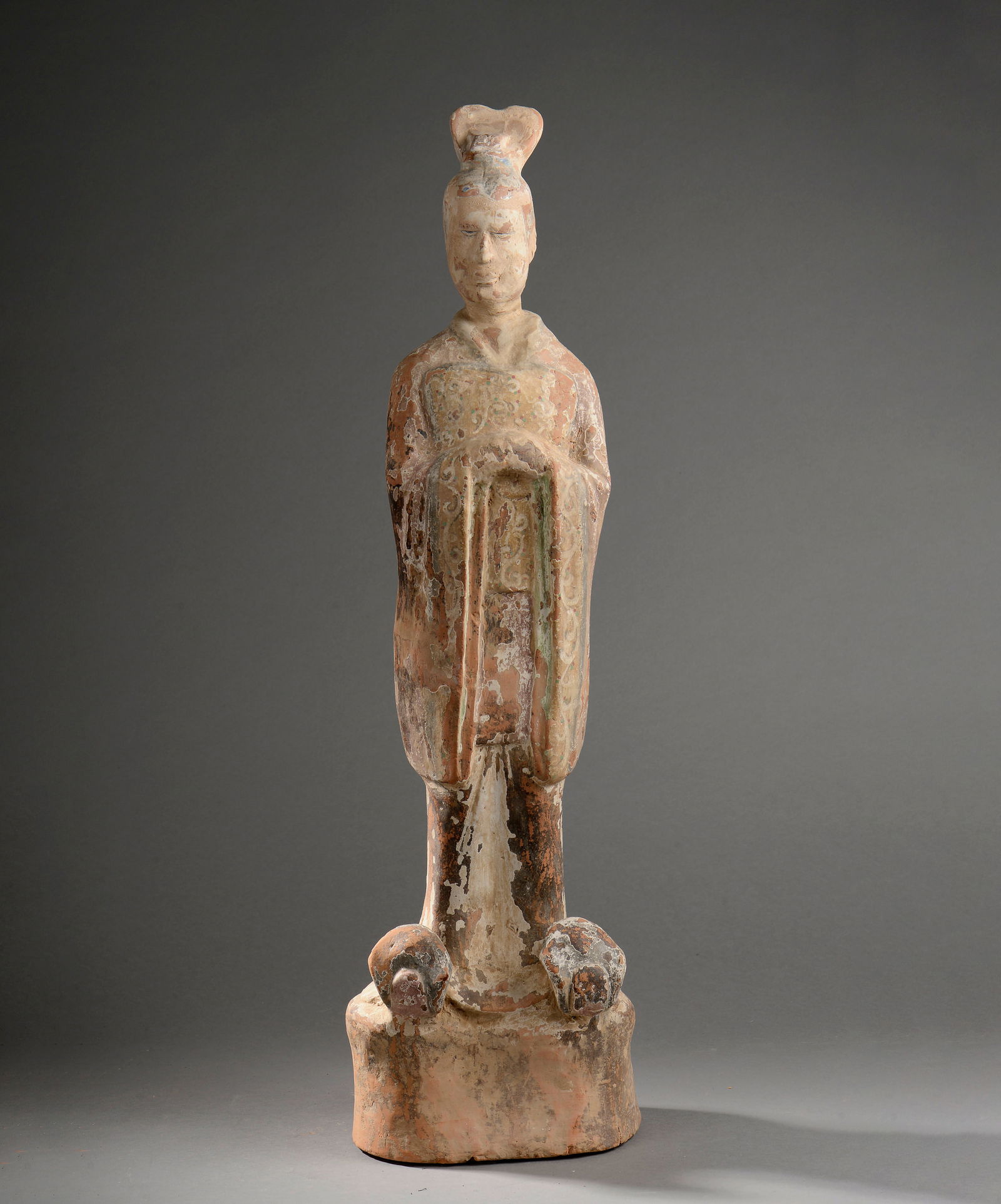 High dignitary standing, Tang Dynasty, China, 7th - 9th century: Terracotta with traces of slip and polychromy. Sculpture depicting a dignitary standing on a mound, dressed in a long tunic over trousers that fall over his shoes, and wearing a high-ranking official'