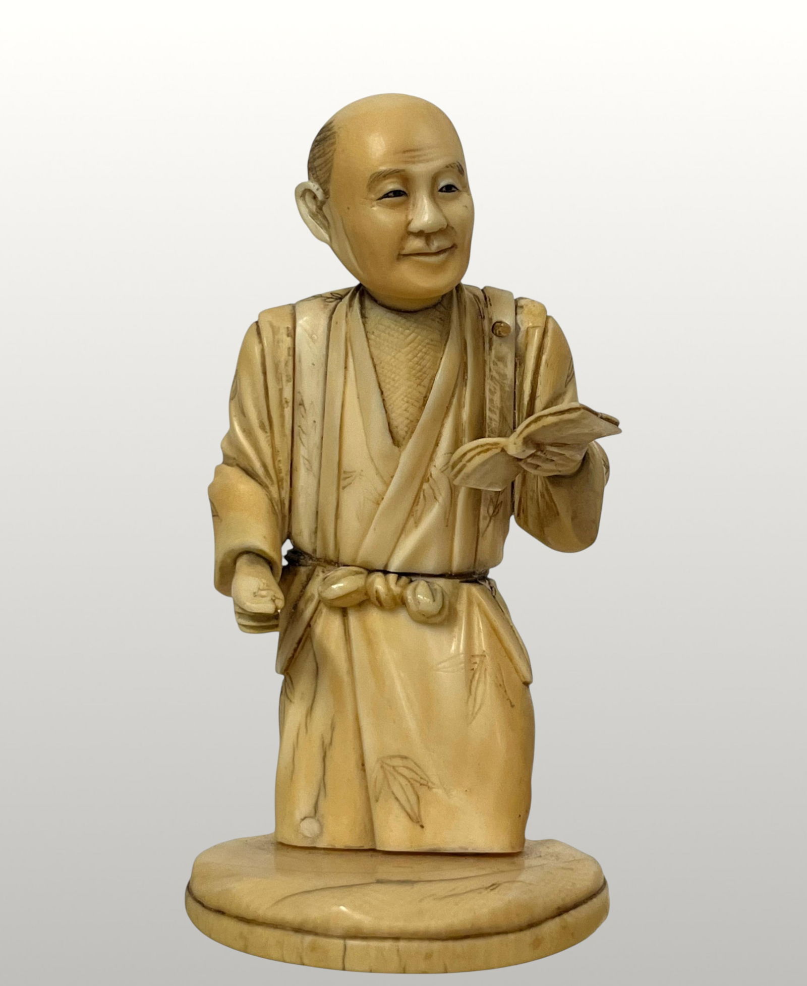 Okimono of a male character with a book, Japan, 19th century: Carved ivory sculpture. Measures 13 cm. Traditional okimono depicting a standing male figure, dressed in a kimono with ample folds decorated with delicate engraved plant motifs. The figure holds a boo