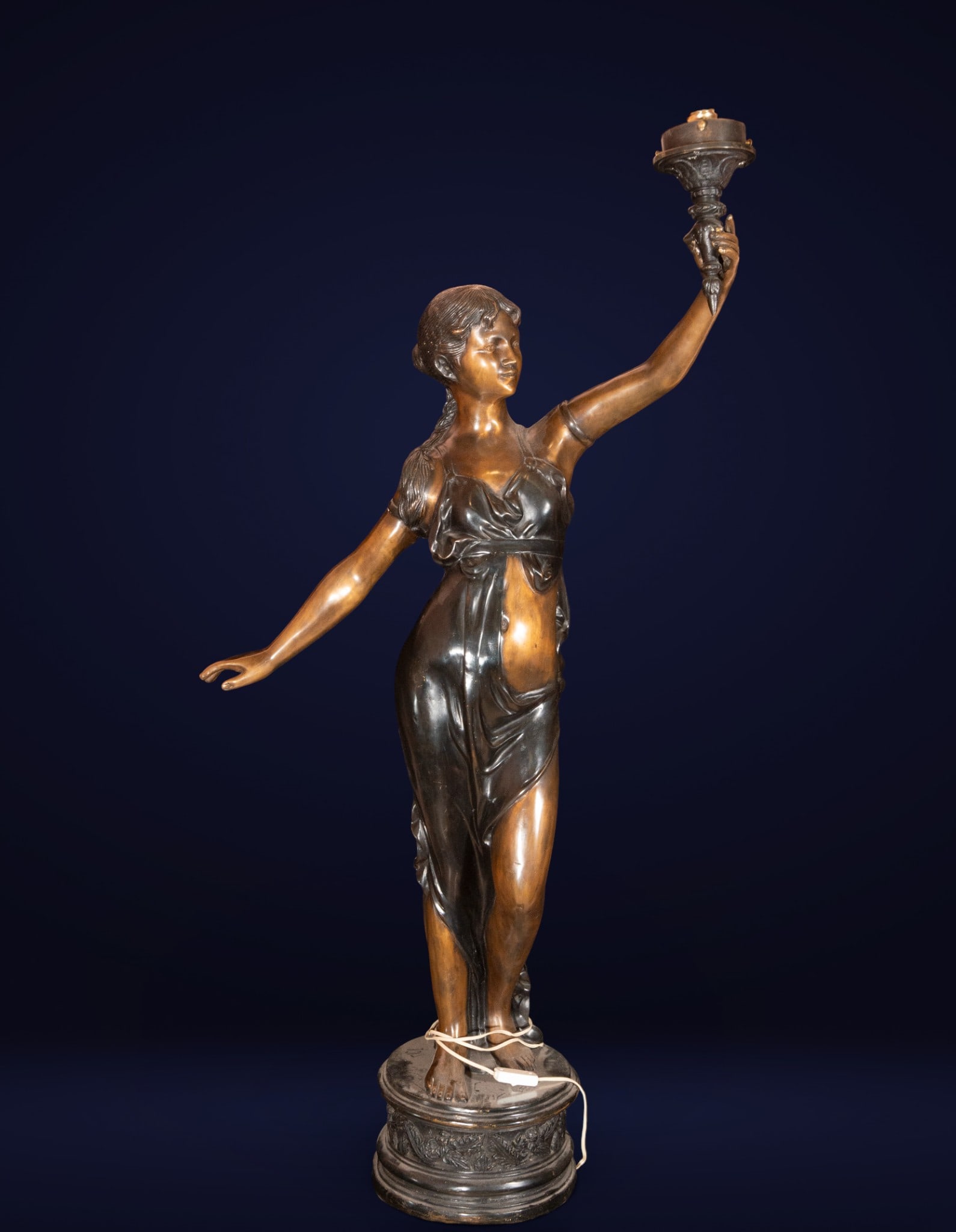 Large electrified bronze torch holder, Beaux Arts style, 1920s: Patinated and partially gilded bronze. Measures 154 x 70 x 30 cm. Standing female figure, conceived with an elegant contrapposto that balances the body's weight on one leg while the other extends gent