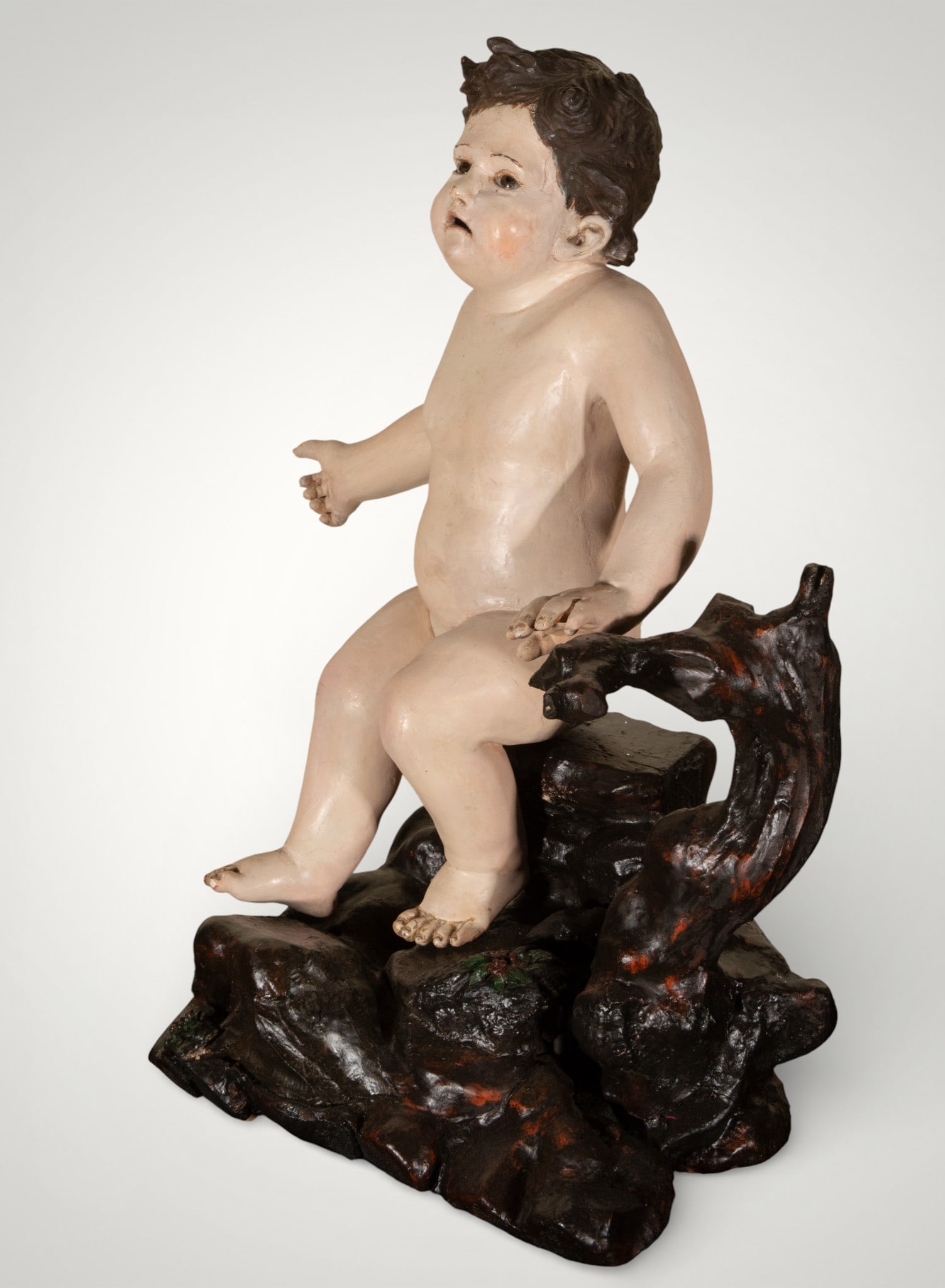 The Seated Boy. Spanish School. 18th Century: Polychrome wood carving. Measures 62 x 47 x 32 cm. This freestanding sculpture depicts a child seated on a naturalistic base. The nude body is modeled with soft volumes and a carefully studied child's