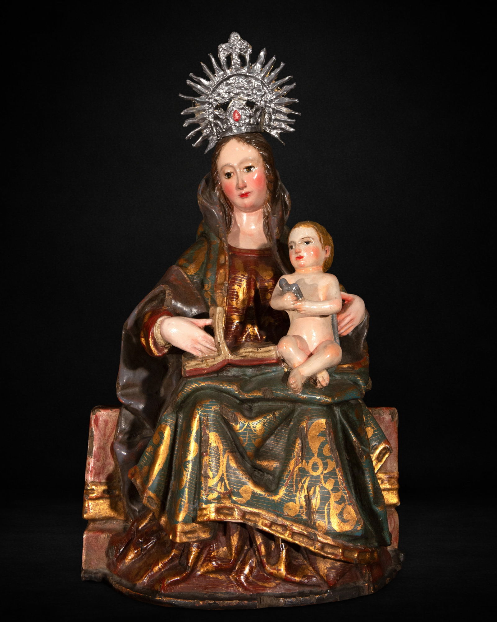 Virgin and Child Seated, 20th Century: Carved and polychromed wood. Measures 42 x 29 x 20 cm. Seated image of the Virgin with the Child on her lap, wearing a crown and metallic halo. Mary holds an open book while the Child, naked, rests na