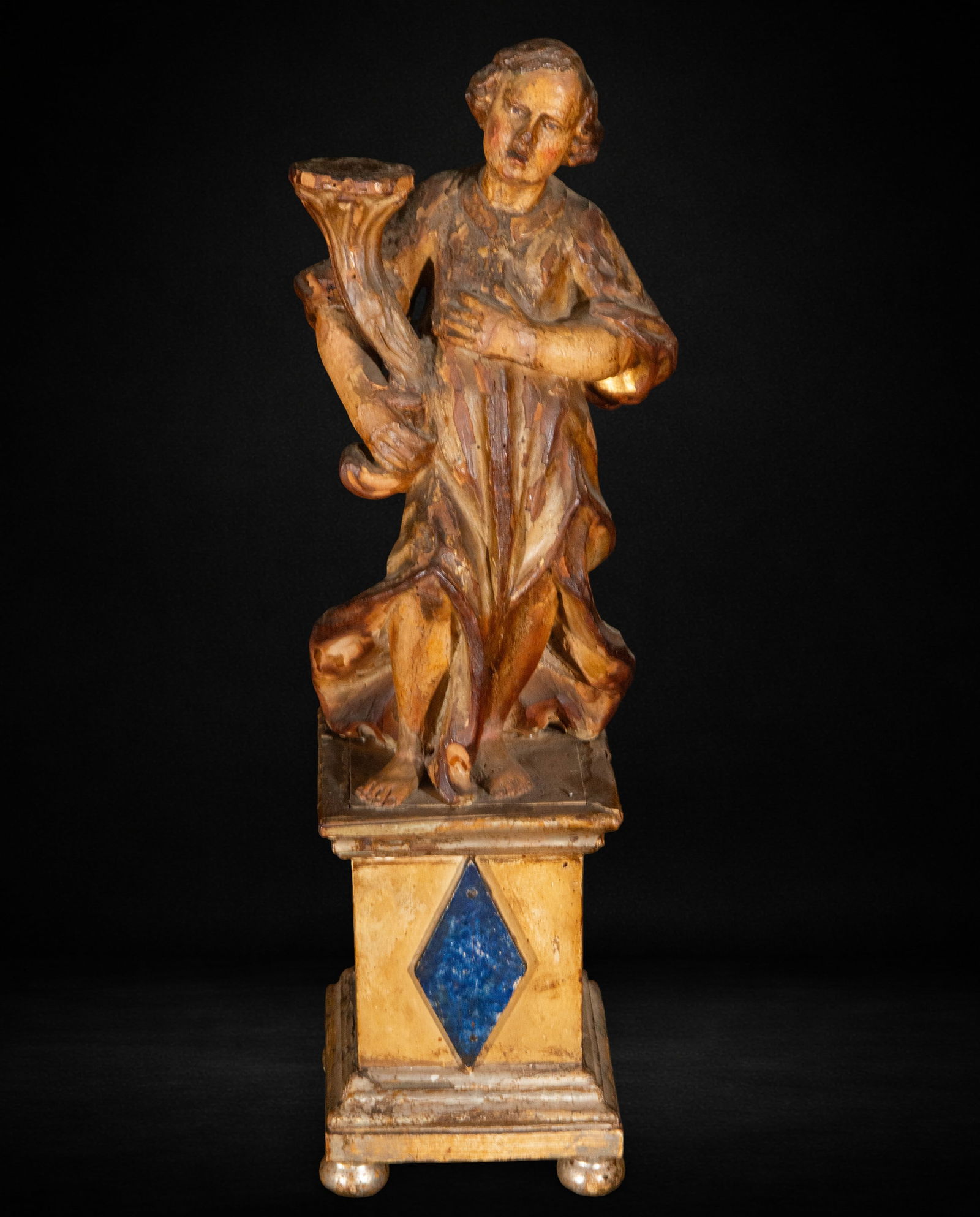 Torchbearer Angel, 17th Century: Carved and polychromed wood. Measures 50 x 13 x 14 cm. Standing torchbearer angel, with a slight twist and a tunic with pronounced folds. He holds a light source. Serene expression on his face, painte