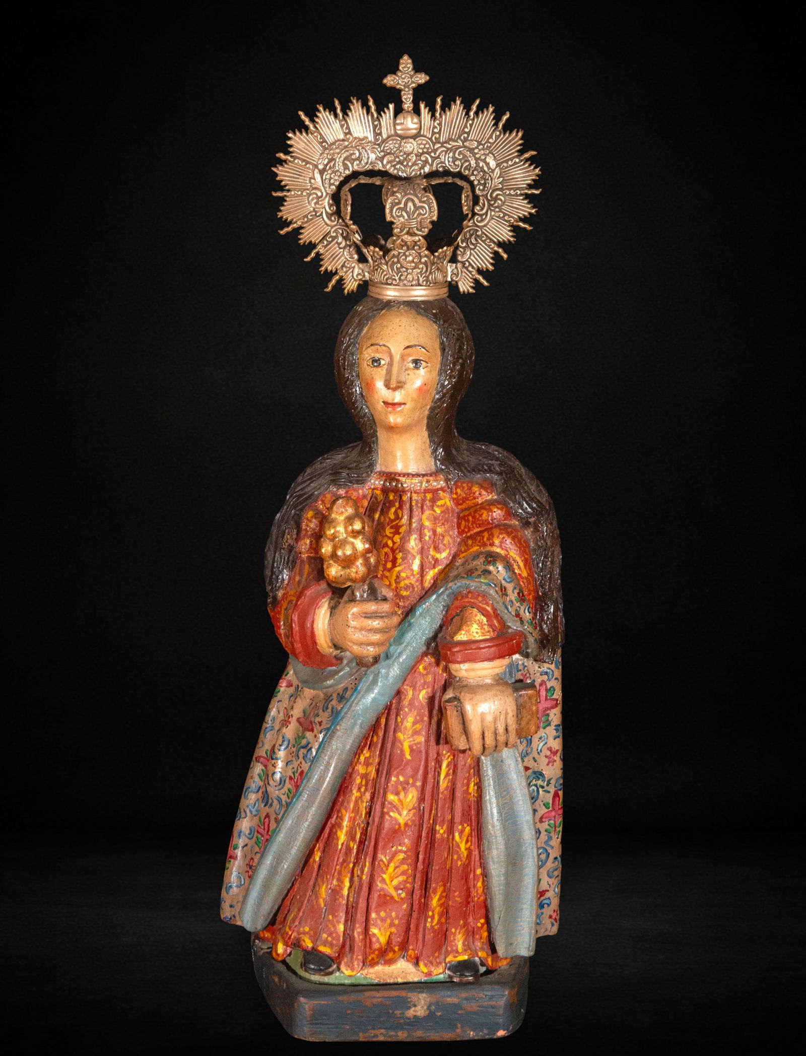 Santa Bárbara, colonial school, 17th century: Carved and polychromed wood. Measures 72 x 27 x 22 cm. Female figure crowned with a metal halo, carrying a palm and a book, dressed in a brocaded tunic in reddish tones and a decorated mantle; frontal