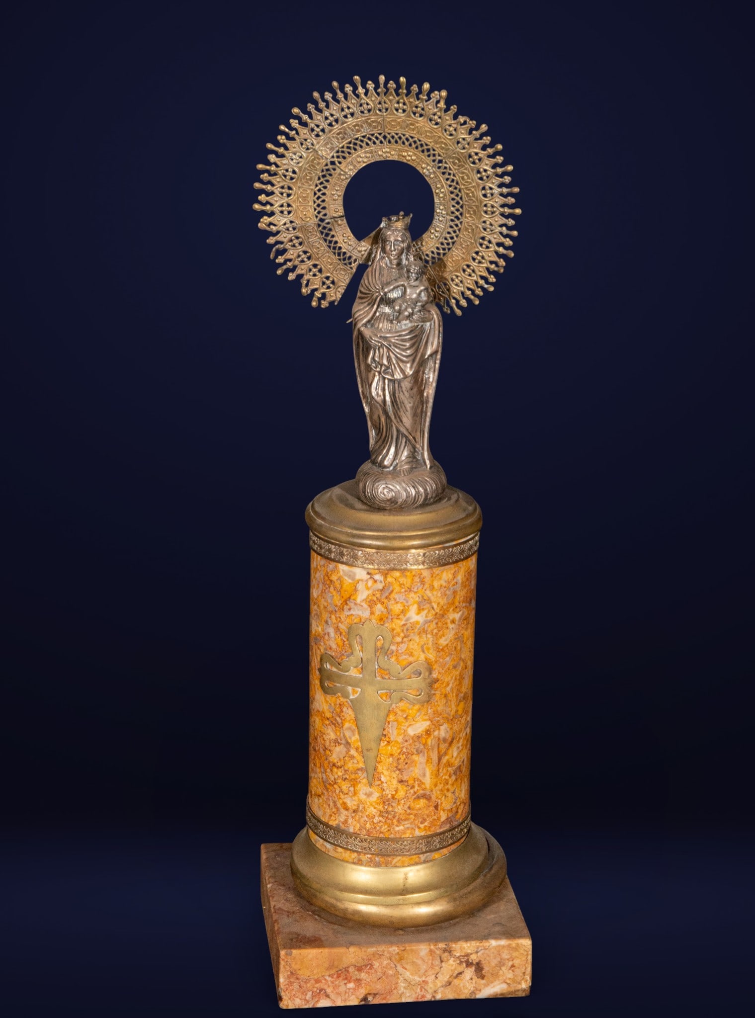 Our Lady of the Pillar, Spanish School, 19th Century: Silver-plated metal and mottled marble. Dimensions: 62 × 17 × 17 cm. Image of the Virgin and Child on a cylindrical column, accompanied by a highly decorative openwork halo and a Cross of St. James