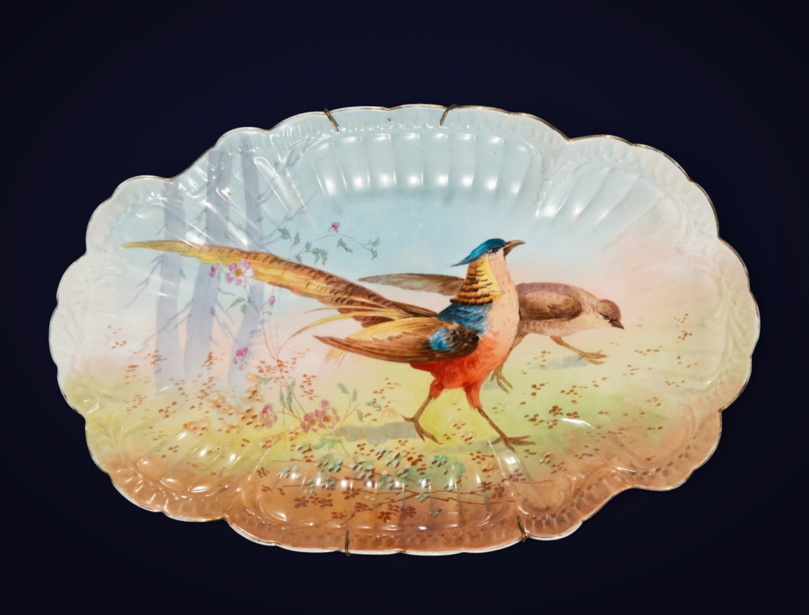 Fruit bowl with birds, European school, 20th century: Earthenware or glazed porcelain. Measures 6 x 47 x 29 cm. A platter with a wavy rim decorated with two long-feathered birds in a soft landscape of flowers and light ground, within a composition of nat