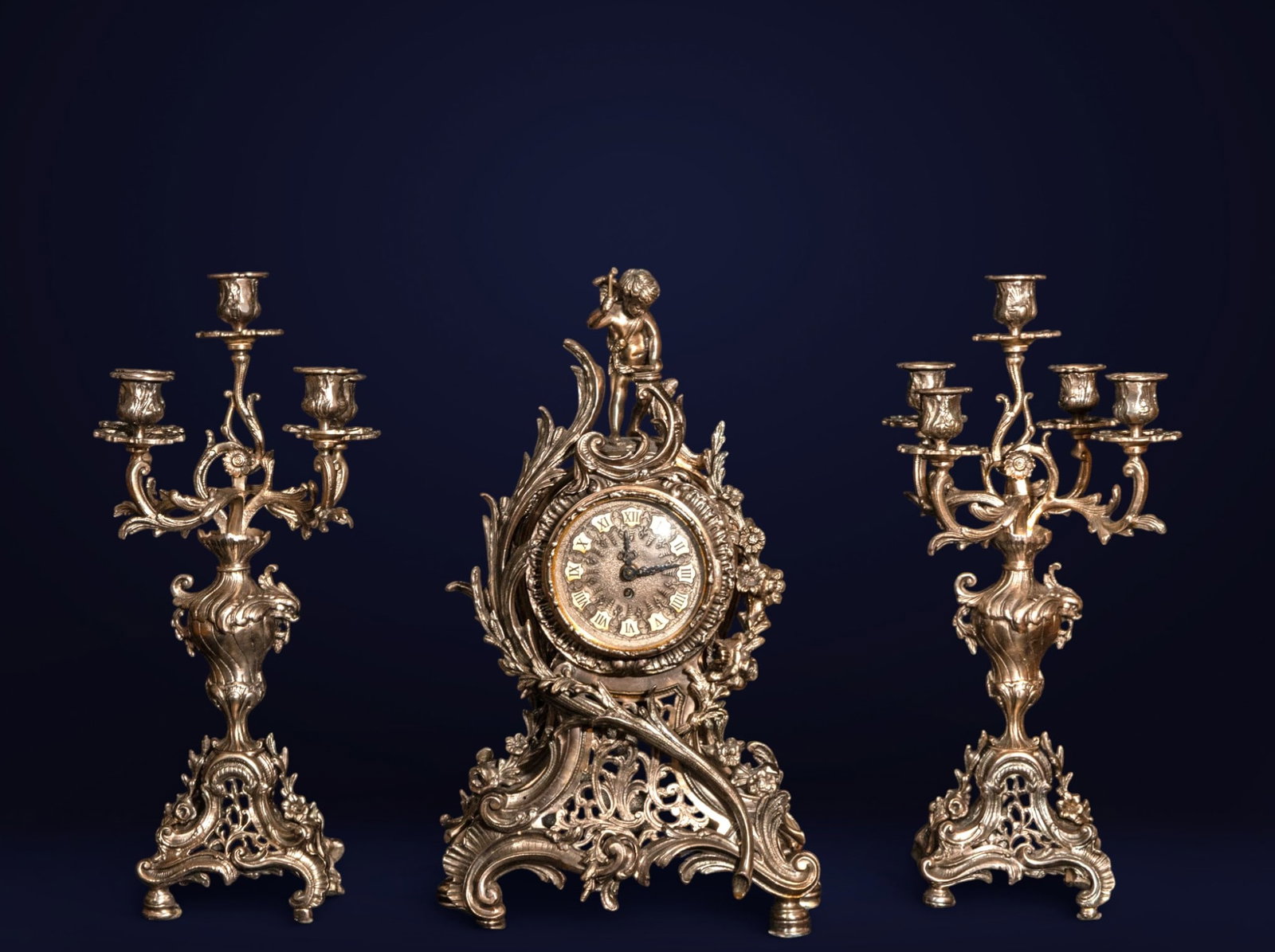 Clock garniture with silver-plated bronze candlesticks, Rococo style, Portugal, 20th century: Silver-plated bronze and metal. Clock measures 55 x 35 x 17 cm; candlesticks 54 x 23 x 21 cm. This decorative set, consisting of a mantel clock and a pair of candlesticks, is designed with an exuberan