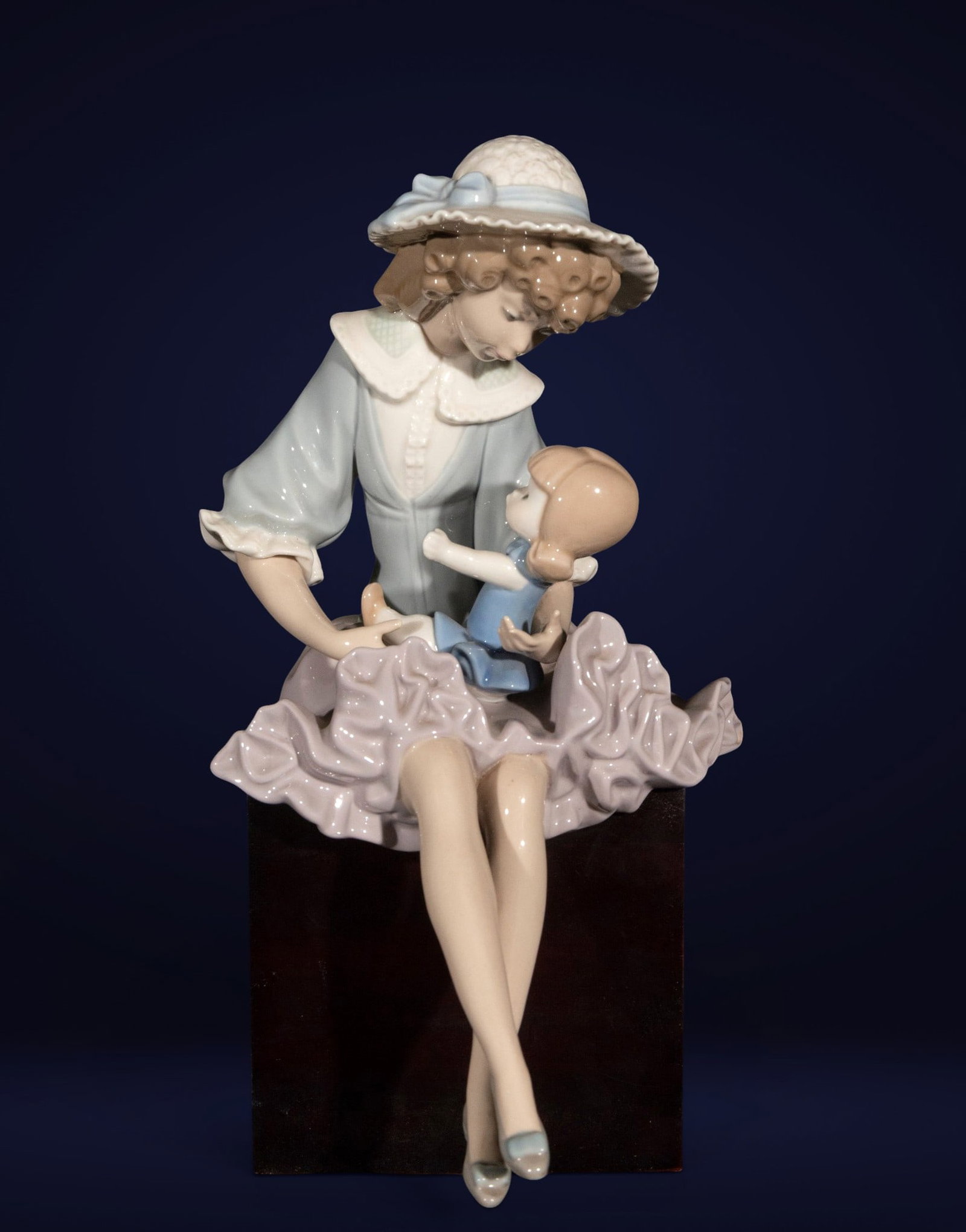 Female figure with child, Spanish School, 20th Century: Glazed porcelain. Dimensions: 17 cm x 16 cm x 20 cm. Seated female figure with child, crafted in soft pastel tones with a glossy finish, featuring delicate modeling of the faces, hat, and folds of the
