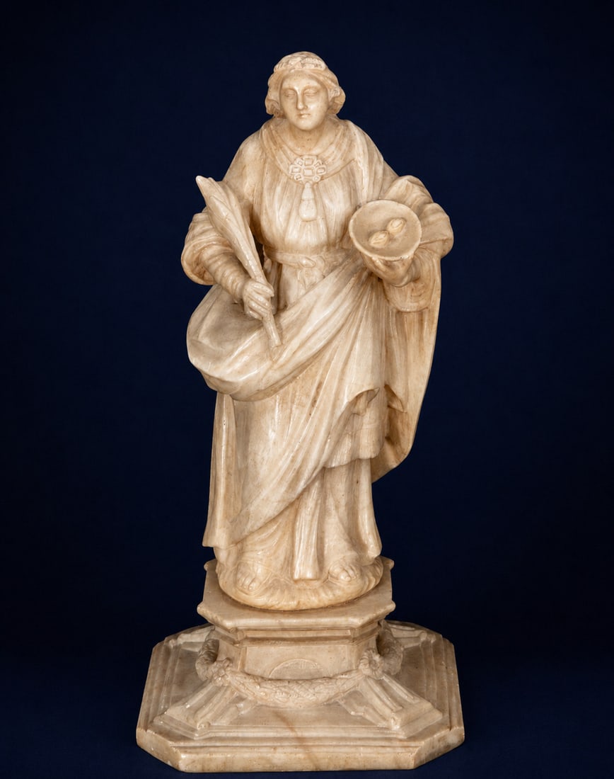 Saint Lucy in alabaster, Italian School, 17th Century: Carved alabaster. Dimensions: 42 x 20 x 17 cm. Sculpture of Saint Lucy depicted standing on an architectural base, holding the palm of martyrdom and a tray with the eyes, traditional iconographic attr