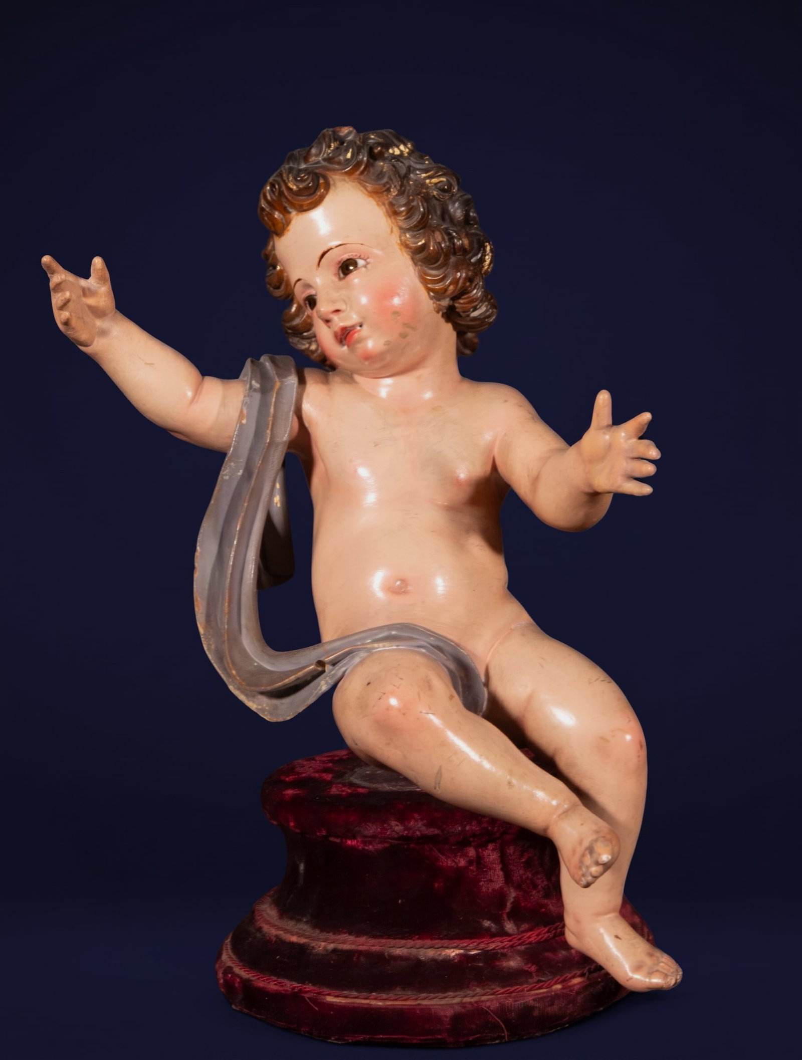 Child Jesus blessing, Spanish School, early 18th century.: Polychrome wood sculpture. Dimensions: 25 x 23 x 21 cm, without base. Freestanding figure of the nude Christ Child, seated on a velvet-covered cylindrical support and partially covered with a bluish-g