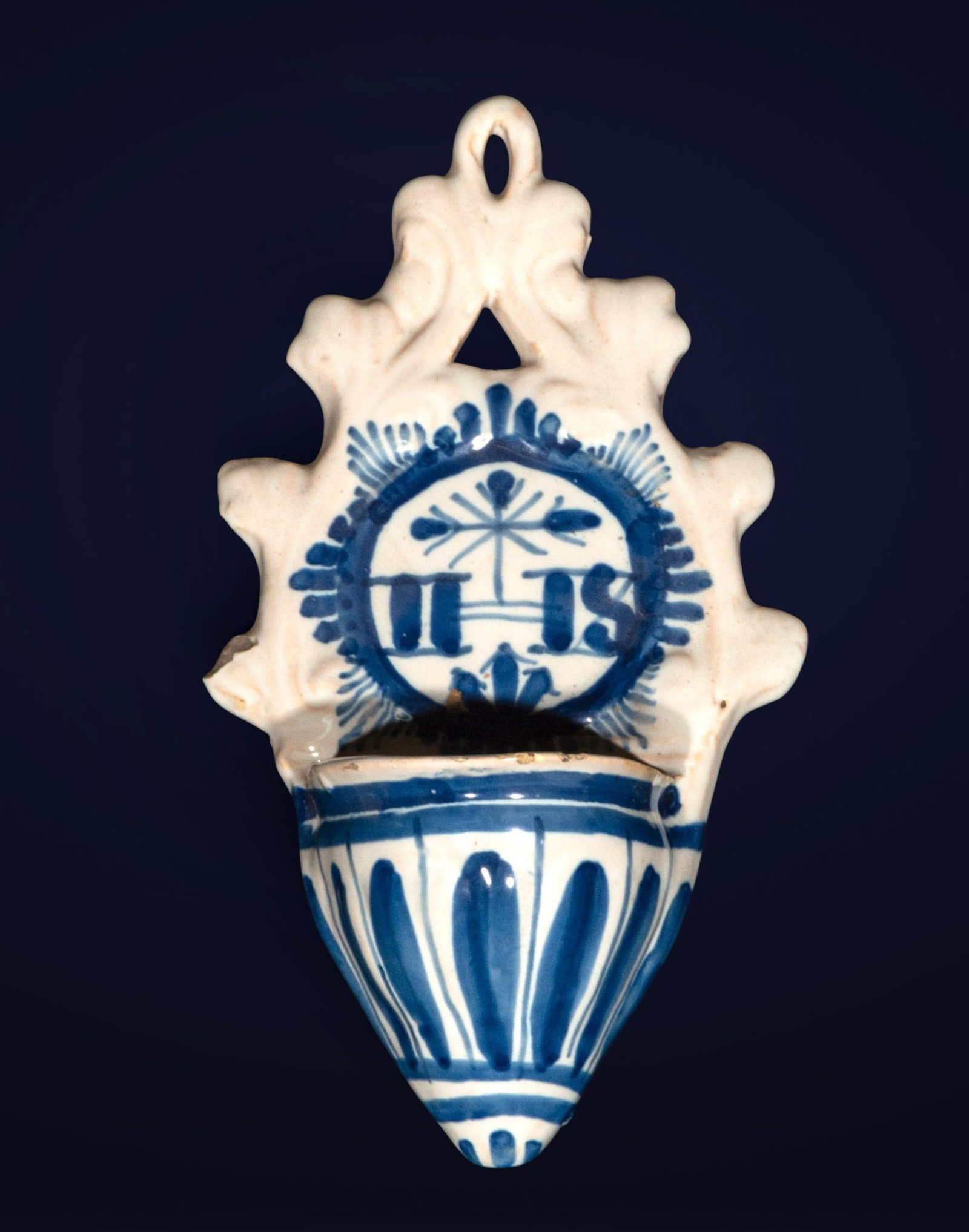 Holy water font of Talavera de la Reina, Castilian School, 18th Century: Glazed and decorated ceramic in blue on white. Measures 24 x 12 x 9 cm. Small wall ewer with a fluted body and a cut-out finial with a top handle, decorated with religious symbols and hand-painted lin