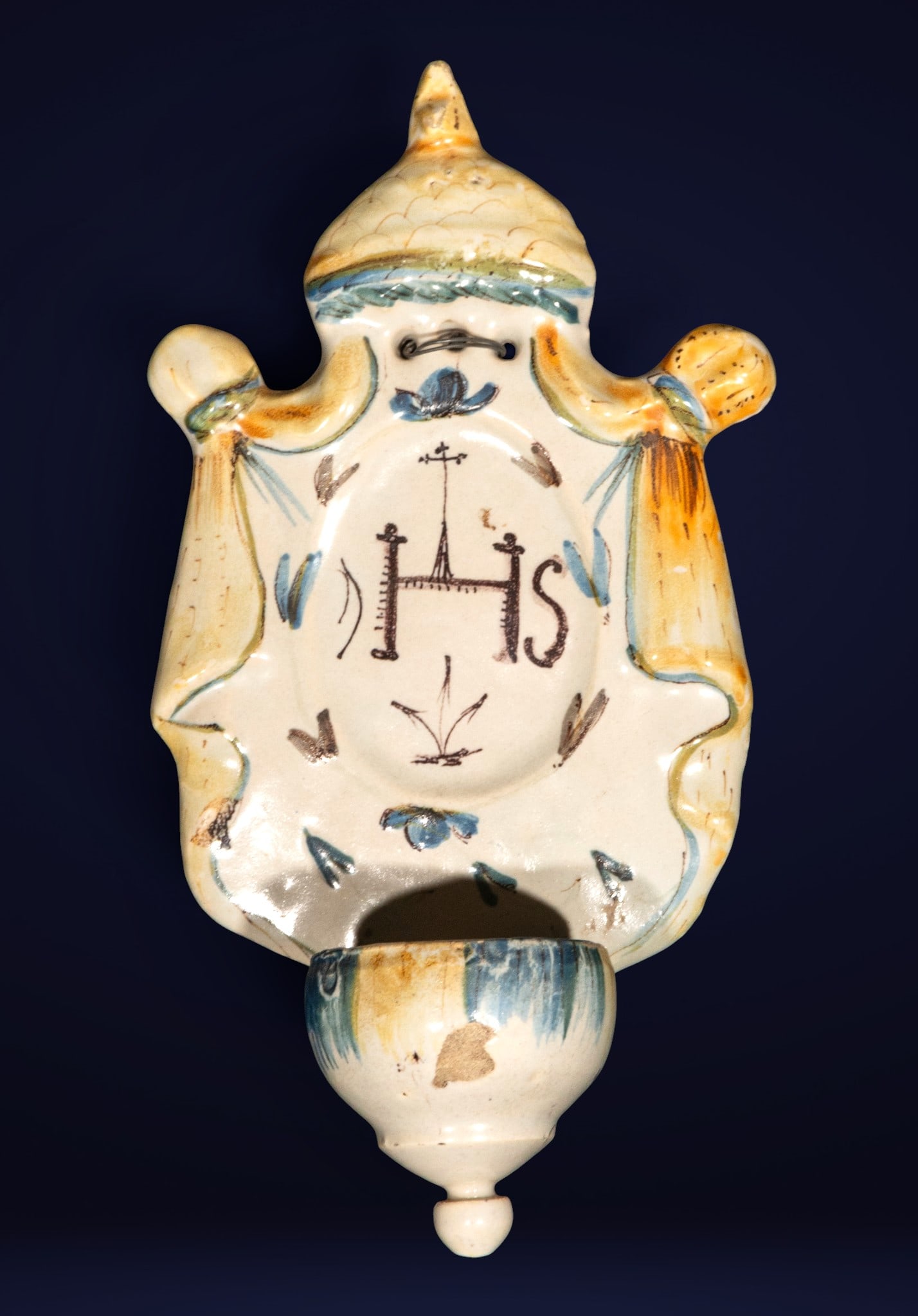 Holy water font with Jesuit coat of arms, Talavera, Castilian School, 18th Century: Glazed and polychrome ceramic. Measures 27 x 15 x 8 cm. Wall-mounted ewer with a mixtilinear profile decorated with the IHS monogram associated with the Society of Jesus, accompanied by floral motifs 