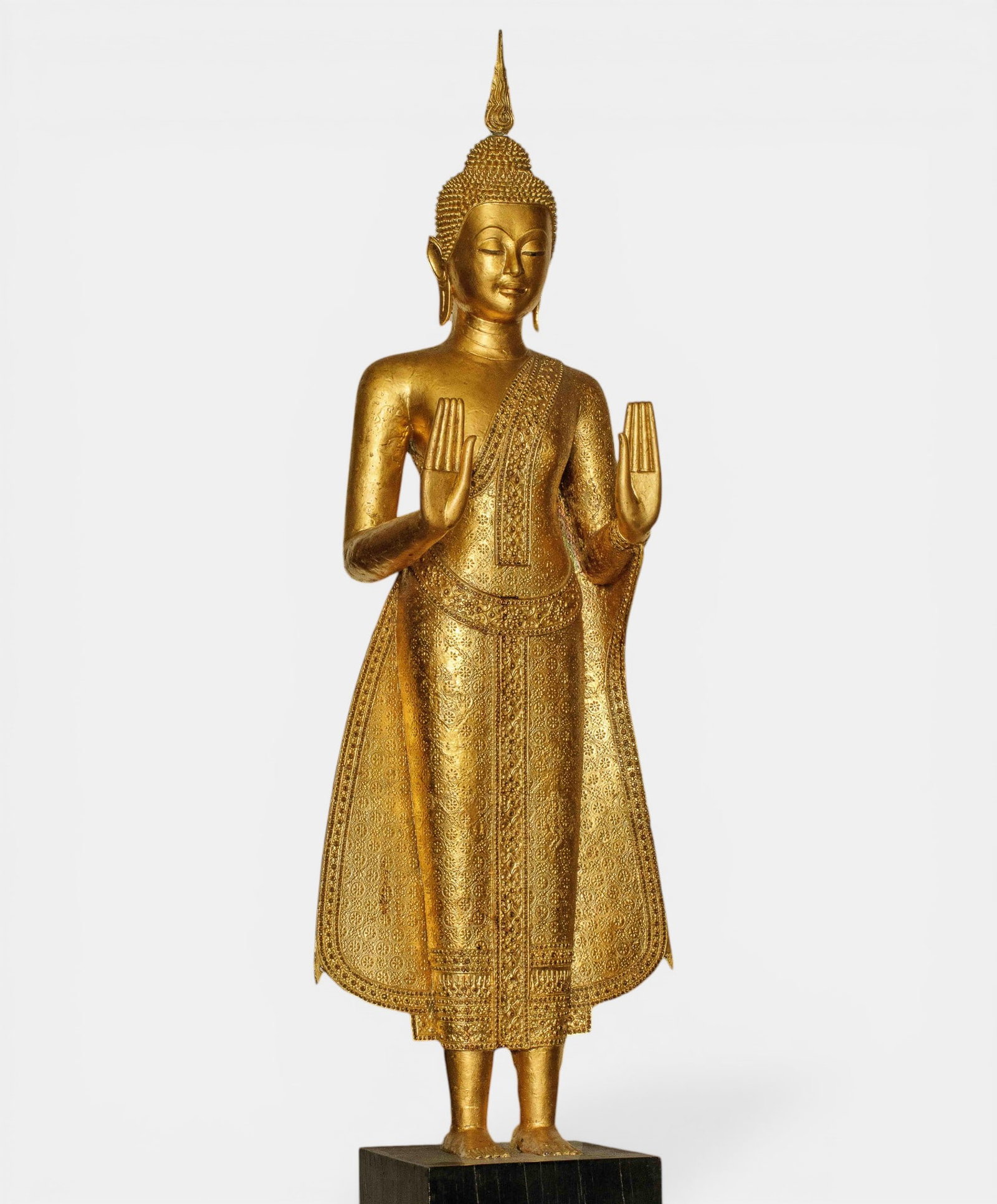 Buddha standing with both hands in abhaya mudra: With a finely chiseled monastic robe and ushnisha surmounted by a prominent rasmi. Gilded bronze. Thailand. 20th century. Buddha: height 120 cm. Wooden base: 62 cm. Total height 162 cm. Lot in interna