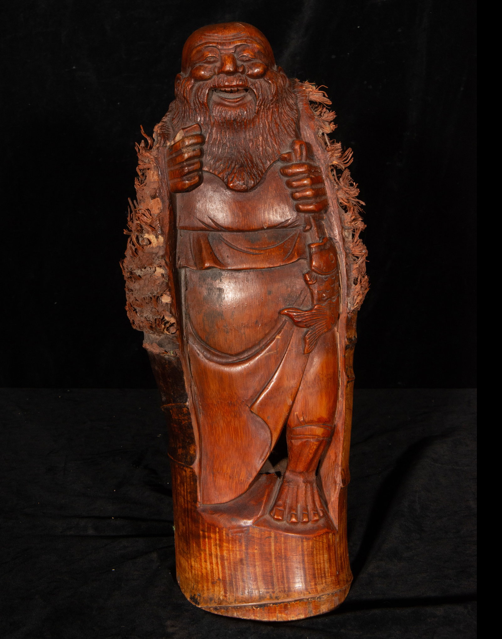 19th Century Chinese School - Sage in Bamboo Root: Carved bamboo. Measures 40 x 18 x 16 cm. Figure of a bearded character with a smiling expression, standing and holding a fish in one hand and a staff in the other. The carving takes advantage of the n