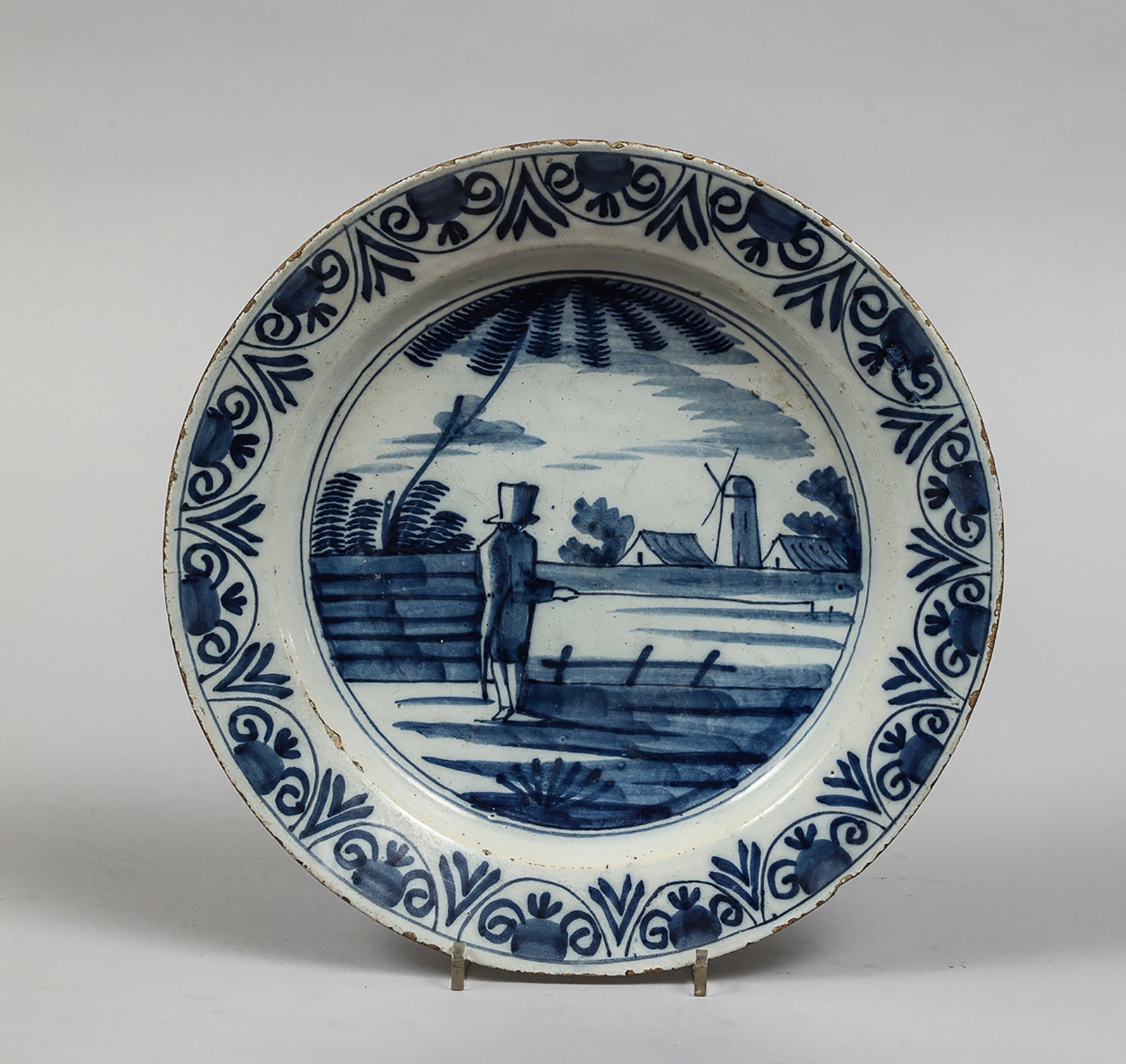Earthenware plate with blue underglaze decoration and figure in rural landscape: The central panel depicts a figure with its back turned in a rural landscape with a mill, trees, and a house. The border is decorated with a stylized vegetal frieze. The reverse is white enamel, unmar