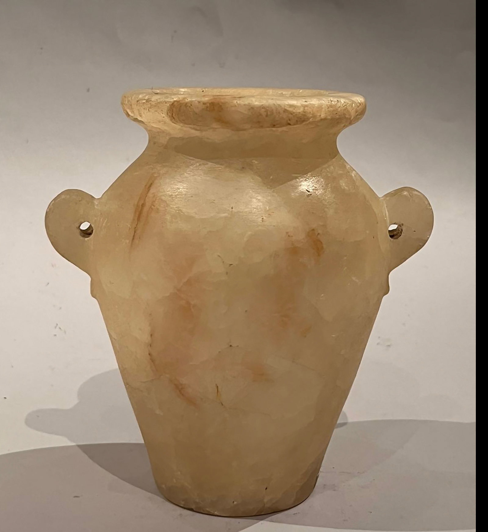 Small Egyptian cosmetic vessel made of alabaster calcite with side handles: A truncated conical vessel, slightly curved and resting on a narrow, rounded base, with an open neck ending in a wide, finely polished, rounded lip. Two small lateral handles on perforated tenons allo