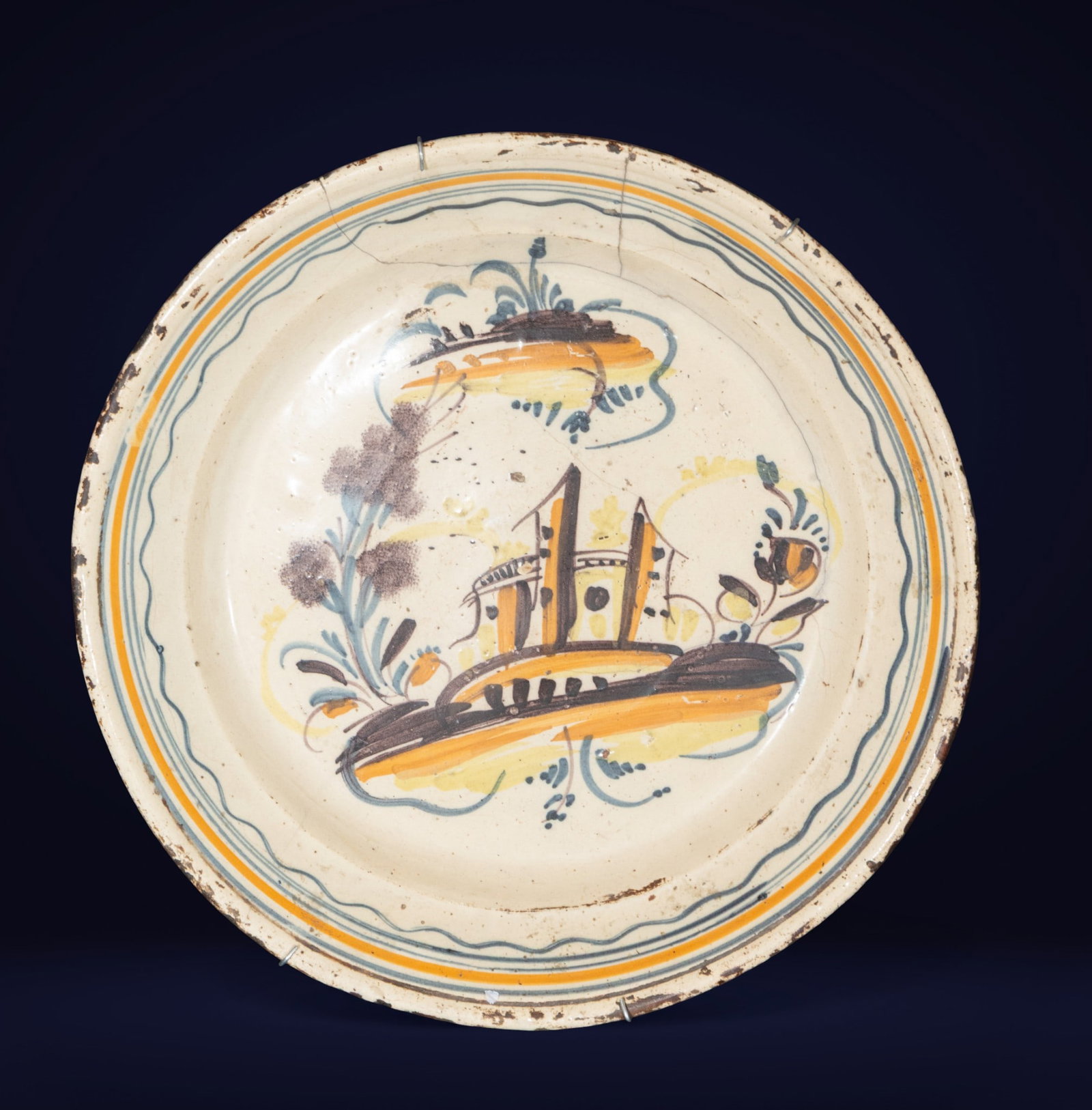 Tricolor plate from Triana. Spanish School. 18th century.: Glazed ceramic decorated in blue, ochre, and brown. Measures 39 x 39 x 7 cm. Architectural and vegetal motifs with loose lines and a wavy border. An outstanding example of 18th-century Triana ceramics