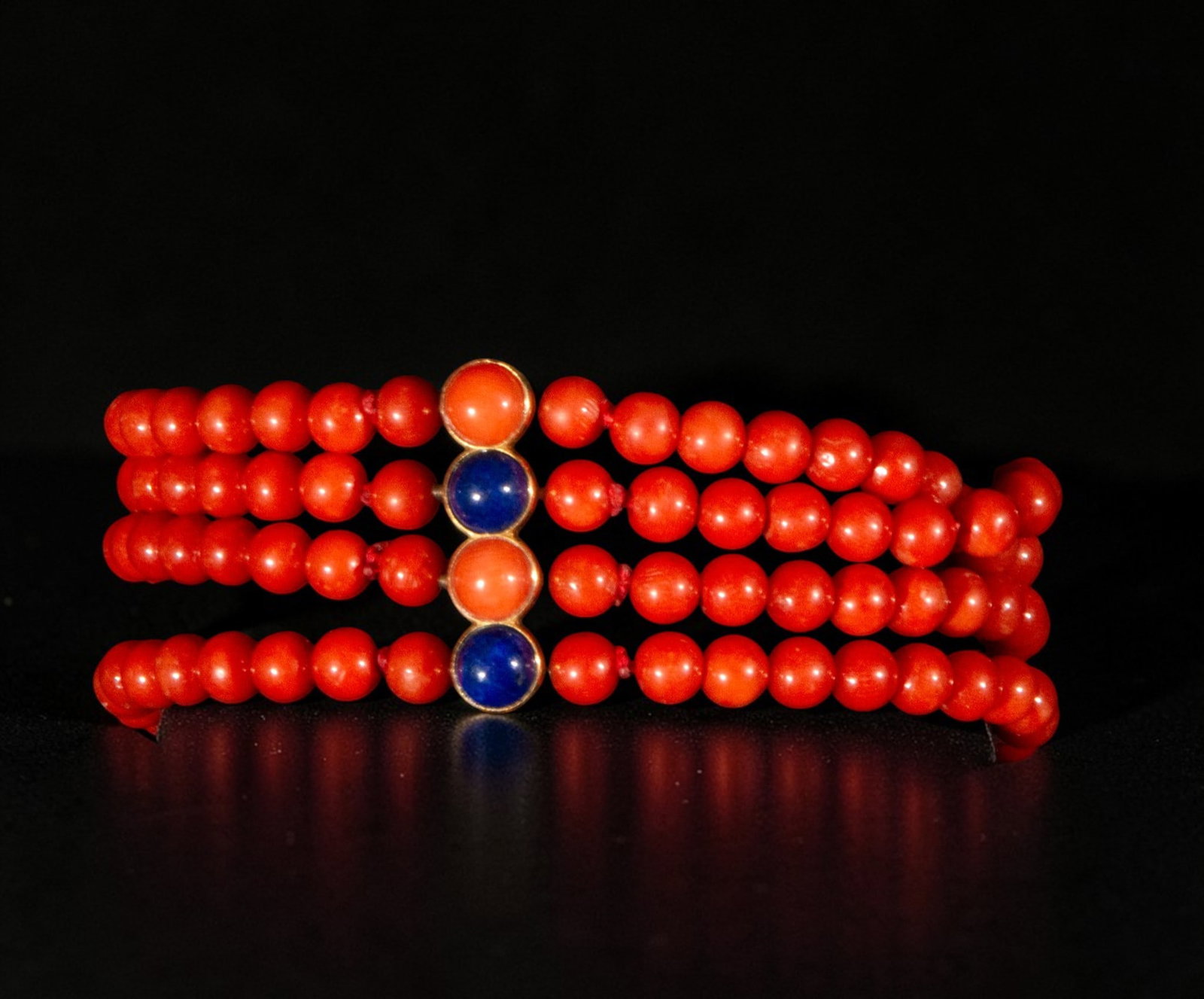 Four-strand coral bracelet with yellow 18k gold clasp and lapis lazuli and coral cabochon: A four-strand coral bracelet with a 18k yellow gold clasp and lapis lazuli and coral cabochons. The design features spherical coral beads arranged in parallel rows, creating a wide, enveloping structu