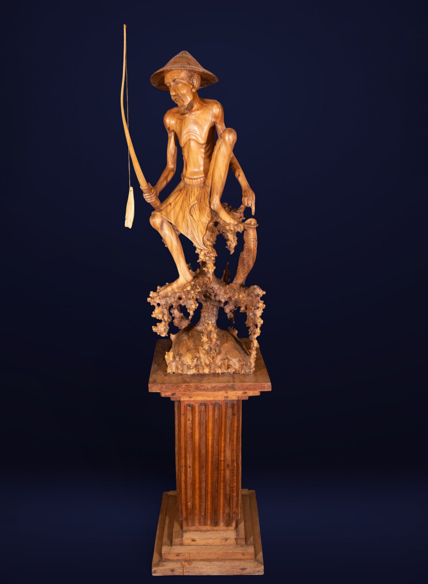 Fisherman on a rock formation, Oriental Art, 20th Century: Wood carving. Dimensions: 142 x 46 x 60 cm. This figurative sculpture depicts an elderly fisherman with a stylized anatomy, wearing a traditional conical hat and holding a fishing rod, standing atop a