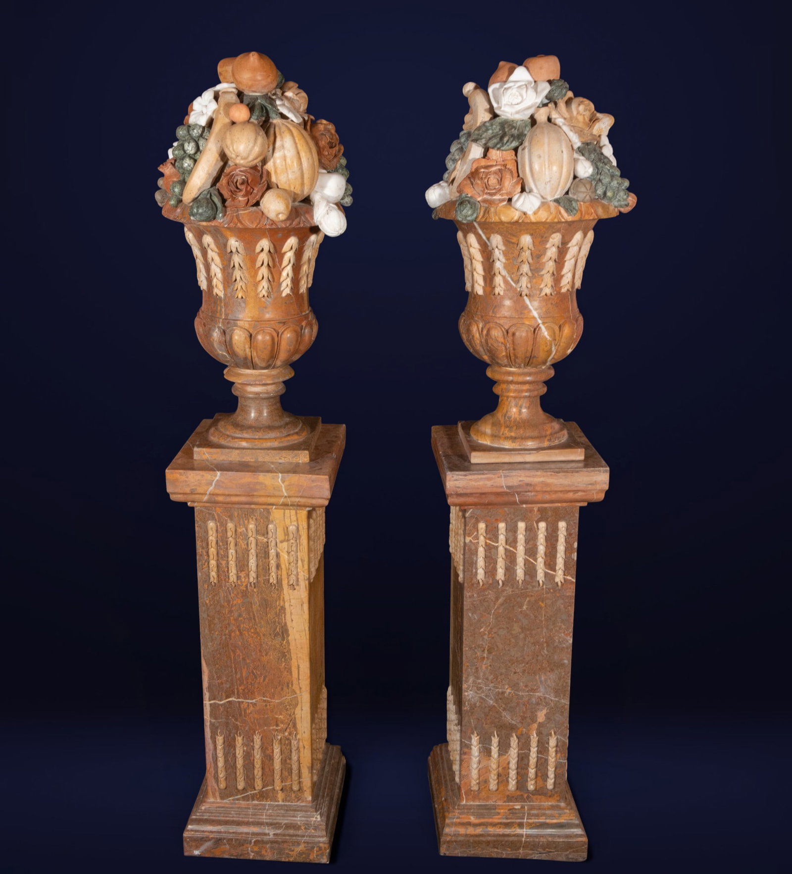 Pair of goblets with garlands of fruit, Neoclassical School, 20th Century: Carved and polychromed marble. A pair of neoclassical-inspired ornamental cups, raised on prismatic pedestals and decorated with relief appliqués evoking vegetal garlands and classical architectural 