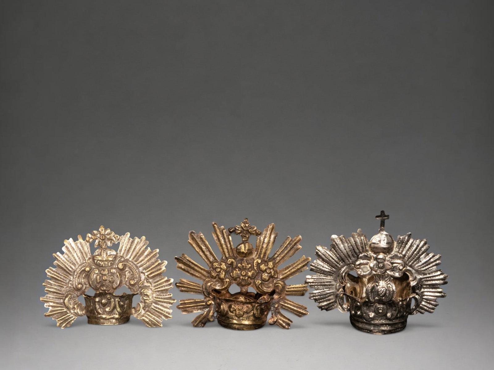 Three alpaca crowns: Set of three embossed alpaca crowns for religious images, with ornamental variations of radiance, flowers, scrolls and cross or cruciferous globe finials, typical of the devotional tradition.