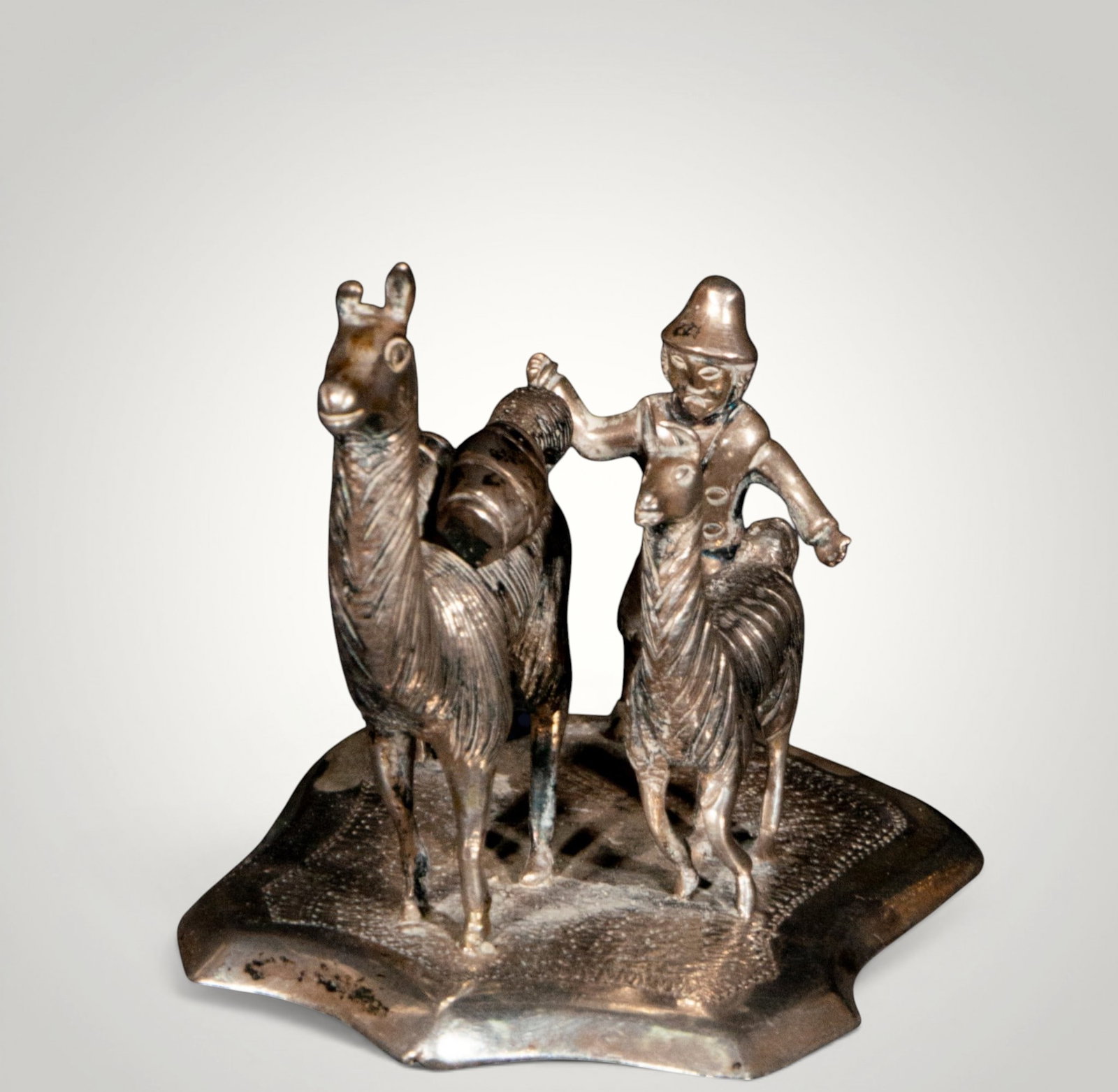 Shepherd with dog and llamas. Peruvian School. 20th Century: Cast, chased, and assembled 800 silver. Measures 8 x 6 cm. Small pastoral scene composed of a standing figure accompanied by a dog and two llamas, arranged on an irregular, organically shaped base. Th