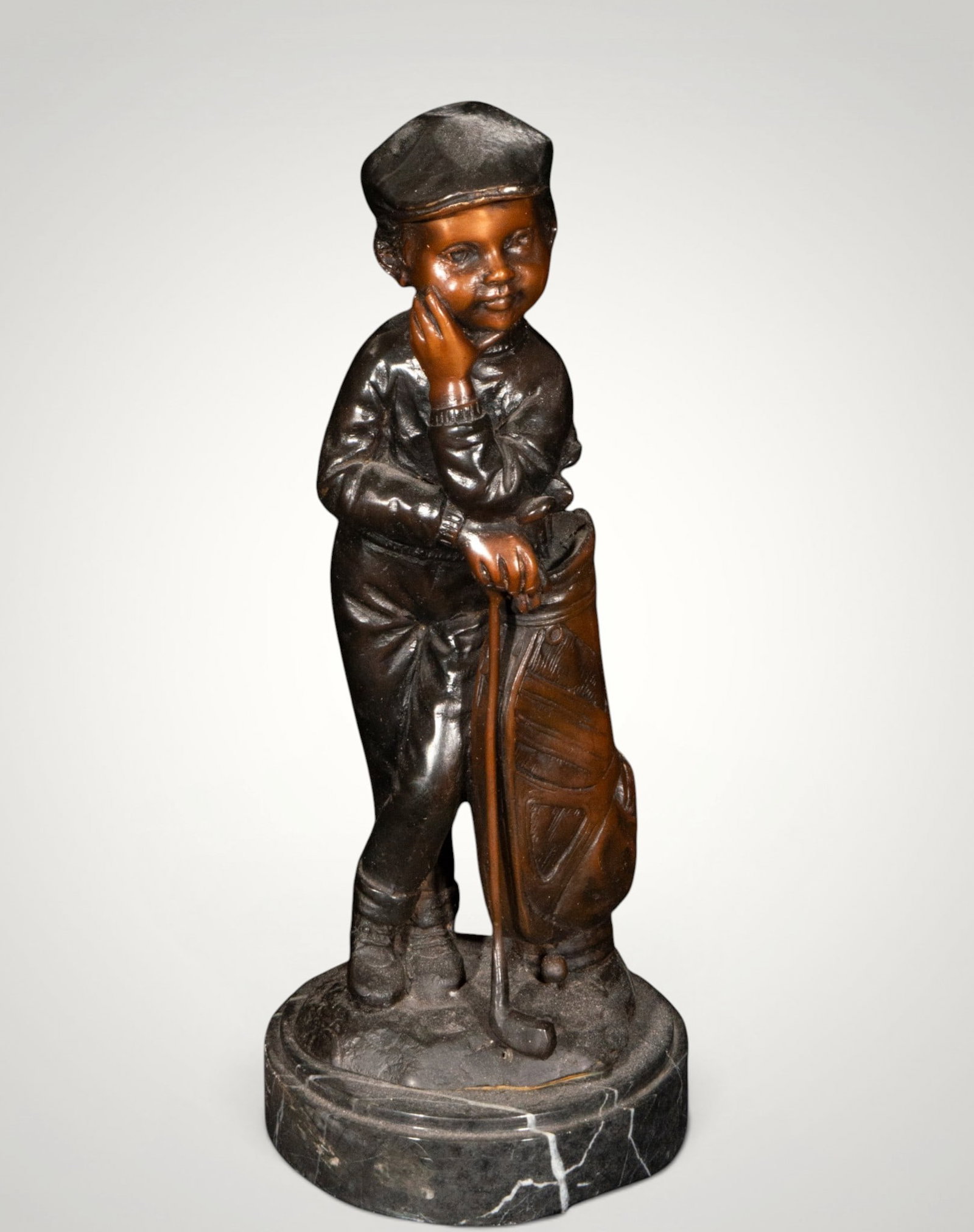 Young golfer. European school. 20th century: Patinated bronze on a marble base. Measures 32 x 13 x 13 cm. A child figure leaning on a golf club next to his bag, depicted in a relaxed and smiling pose. The naturalistic modeling, the details of th