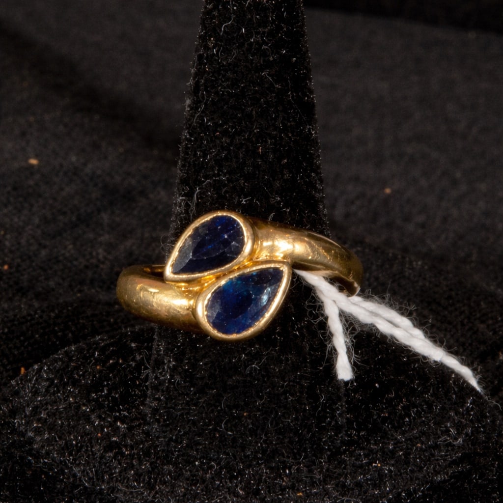 18k you and me sapphire ring: A gold-tone 18k gold ring with an open, toe-and-me design, featuring a smooth, voluminous band that culminates in two pear-shaped blue sapphires, facing each other in a symmetrical arrangement. 