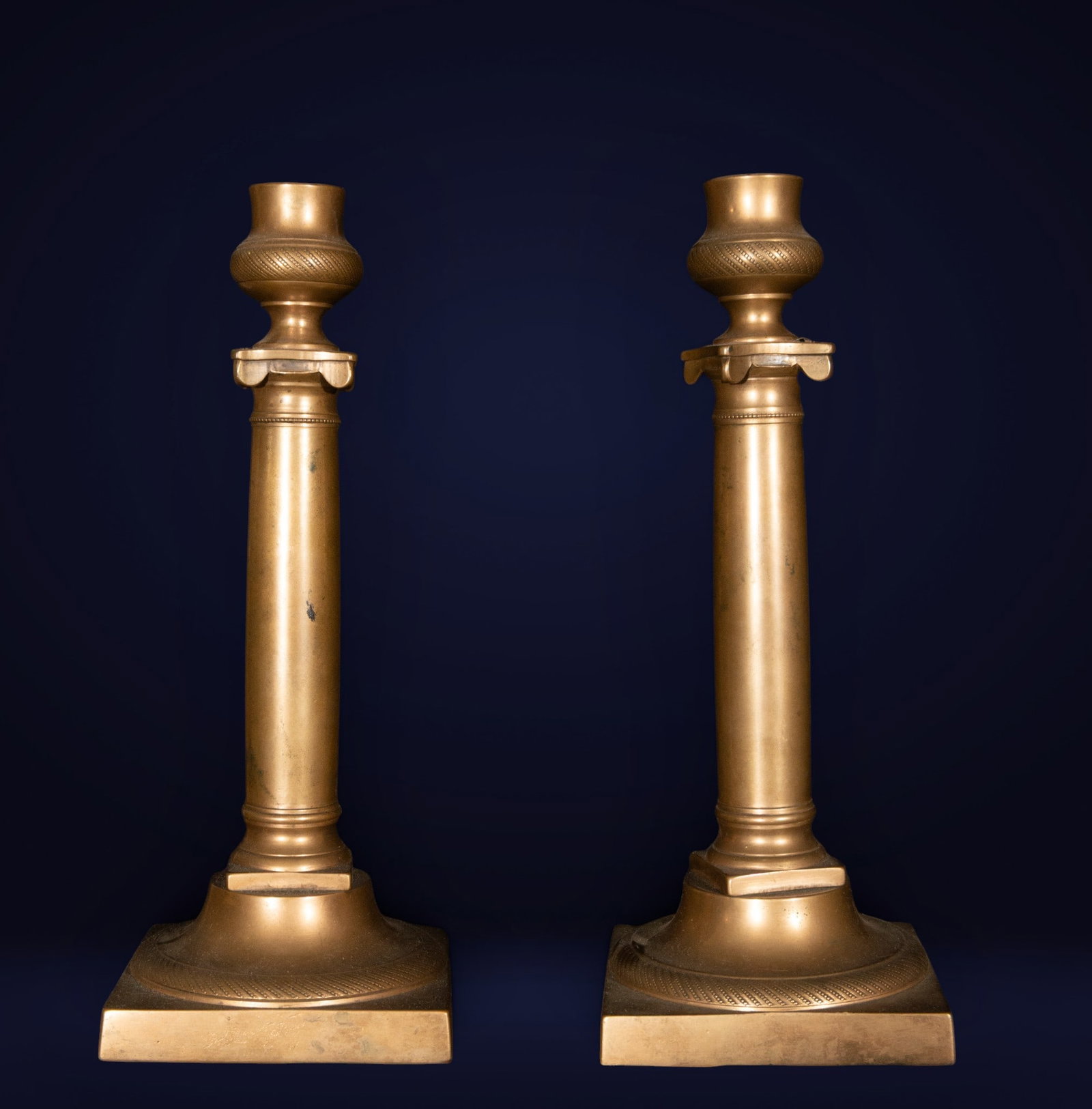 Pair of Empire bronze candlesticks, French School, 19th century: Gilded and patinated bronze. Dimensions: 27 x 11 x 11 cm. Pair of single-arm candelabra in the Empire style, with an architectural structure of clean lines and balanced geometric proportions. The cyli