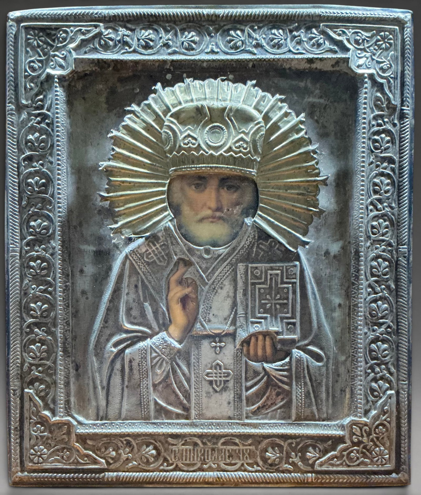 Icon of Saint Nicholas of Myra with oklad, Russian School, 19th Century: Metal front and cloth back. Measures 31.3 x 26.3 cm. Icon with a frontal bust of Saint Nicholas of Myra covered with a rich, chased metal overlay featuring vegetal and geometric motifs. The saint hold