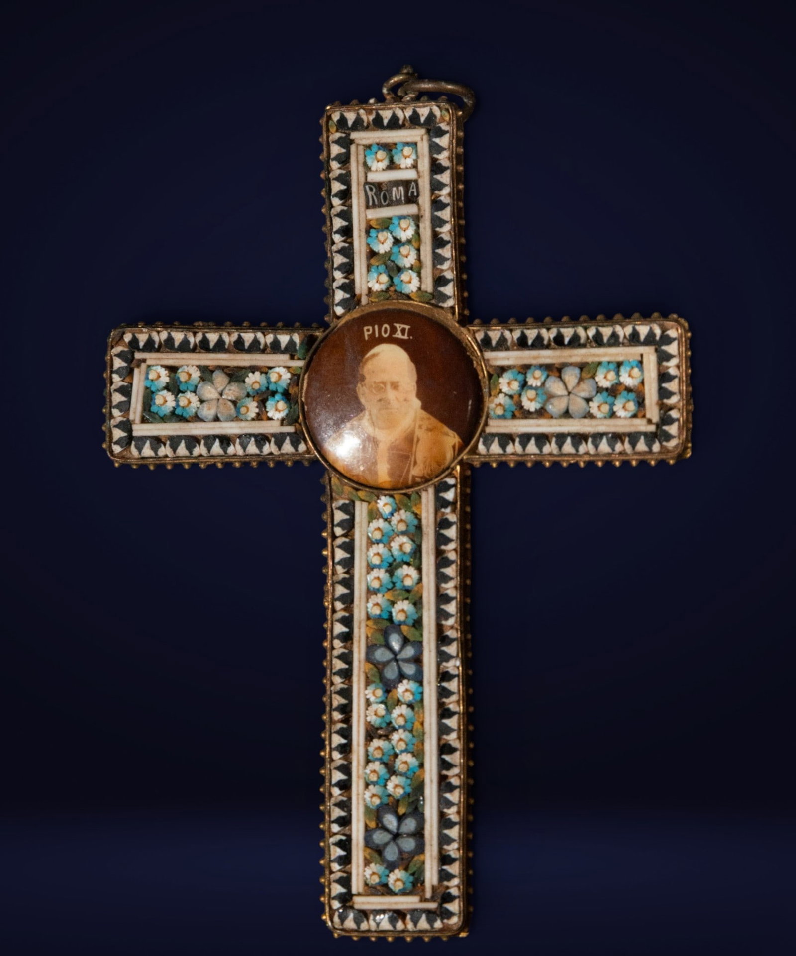 Grand Tour micromosaic cross with portrait of Pius XI, Italian School, 19th Century: Micromosaic and metal. Dimensions: 9 cm x 6 cm x 0.5 cm. Decorative cross crafted with delicate floral micromosaic work in vibrant shades of blue, white, and black, centered by a circular portrait of 