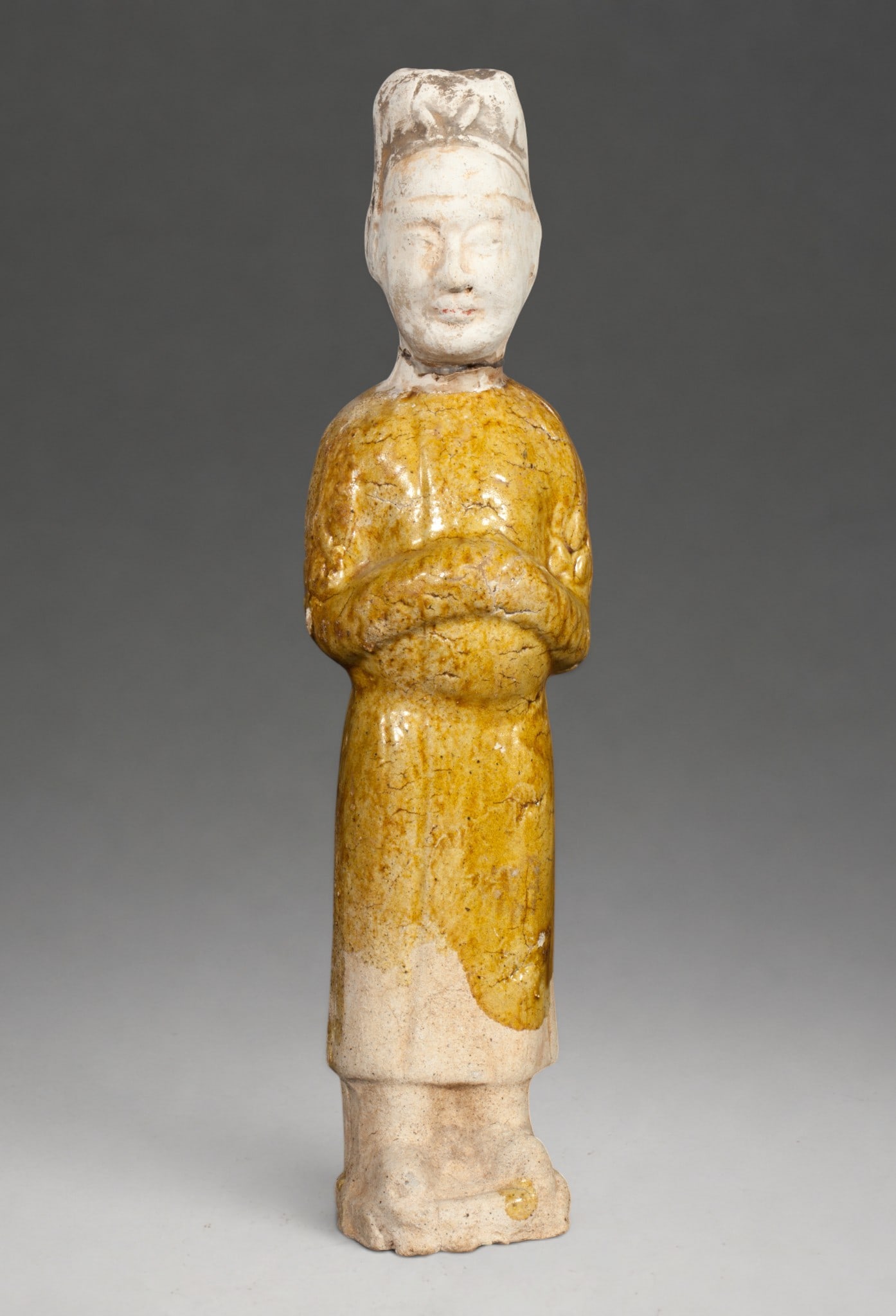 Funeral dignitary in robe, Tang Chinese school, 7th-9th century: Partially glazed terracotta. Measures 23.5 cm in height. Funerary figure of a dignitary depicted standing with hands crossed over the chest, dressed in a long ceremonial robe and a tall, official head