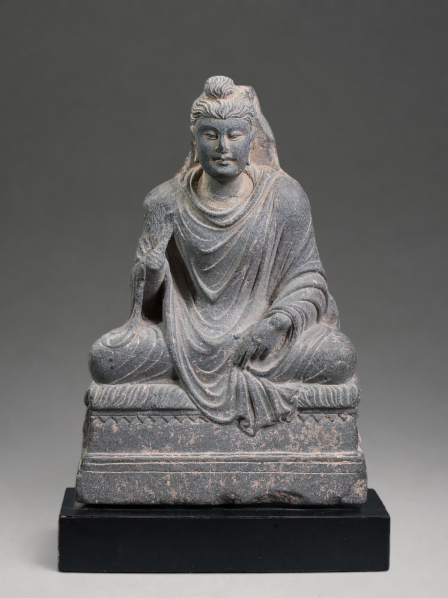 Exceptional Maitreya Gandhara Buddha, 1st-3rd Century: Carved gray schist. Measures 21 x 14 cm. Sculpture of Buddha Maitreya seated on a quadrangular base, with crossed legs and dressed in a monastic robe with deep folds. The face has a serene expression,