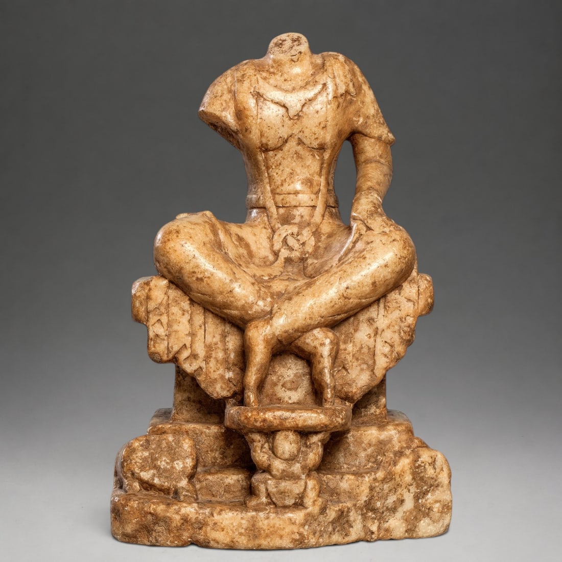7th Century Chinese Buddha: Carved white marble. Measures 29 x 17 x 15 cm. Headless sculpture of a seated Buddha on an architectural throne with crossed legs and feet resting on a support held by a human figure. The body is enve