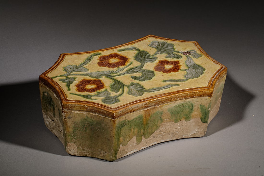 Sancai funeral pillow with floral decoration, Chinese school, 19th - 20th century: Stoneware with a Sancai glaze in shades of green, yellow, and brown. Measures 10 x 29 x 19 cm. Rectangular funerary pillow with slashed sides and a top surface decorated with stylized flowers and stem