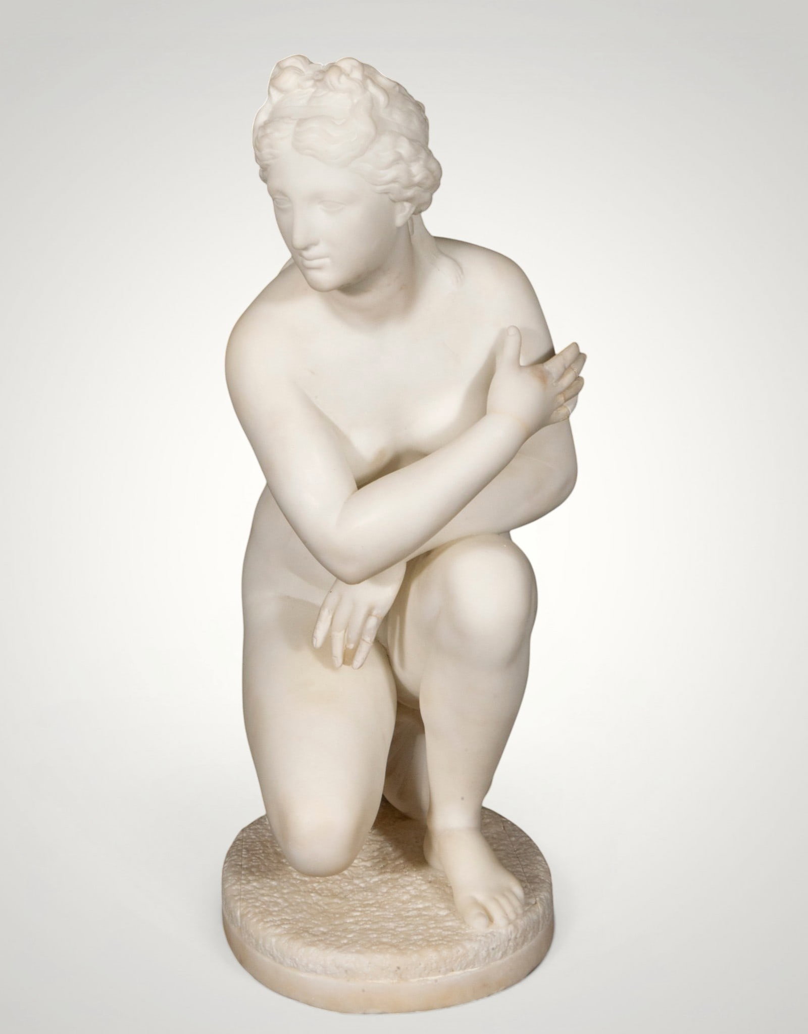 Venus in marble. European School. 19th century: Carved white marble. Measures 52 x 23 x 27 cm. Nude female figure in a crouching position, with her arms crossed over her torso and her head gently inclined. The idealized anatomy, the uniform polish,