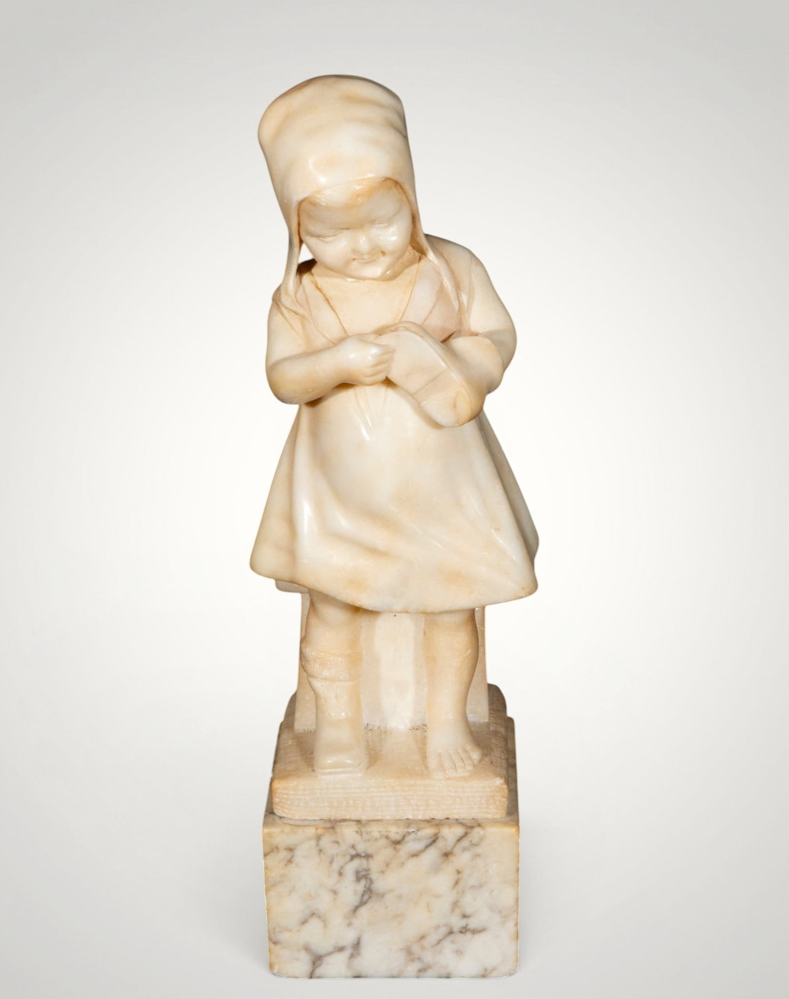 Girl with clogs. European school. 19th century: Carved alabaster. Measures 32 x 12 x 9 cm. Standing figure of a child wearing a cap and simple dress, focused on the clog held in its hands. The gentle modeling, polished surface, and anecdotal charac