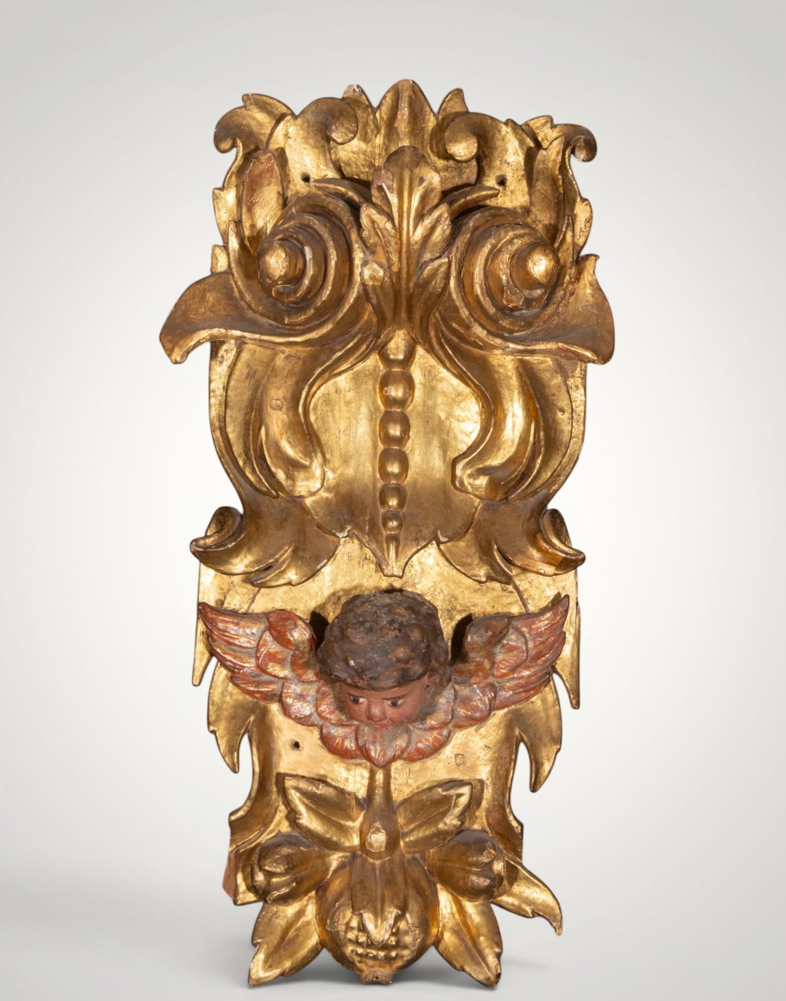 Ornamental appliqué with angel's head. Hispanic School. 18th century: Carved, polychromed, and gilded wood. Measures 93 x 47 x 14 cm. Vertical decorative element with a winged angel's head, scrolls, leaves, and floral motifs arranged on a gold background. The relief car