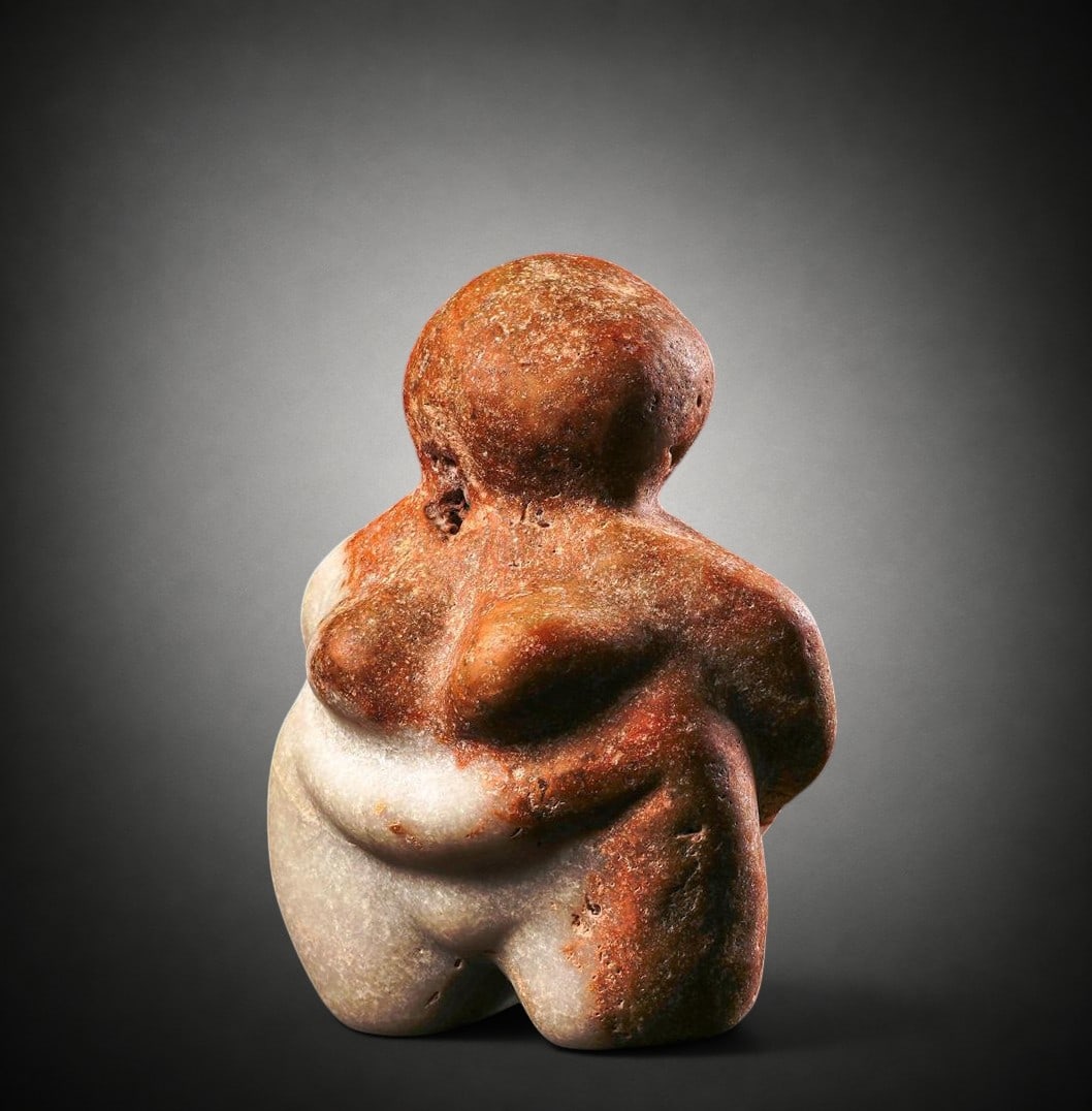 Neolithic steatopygic female figurine in two-tone marble, Mediterranean region, c. 5000–3000 BC.: White marble and orange-brown marble, carved from a single piece with natural veining and inclusions. Measurements: approximately 9.5 cm in height. Weight: approximately 385 g. Neolithic female figure