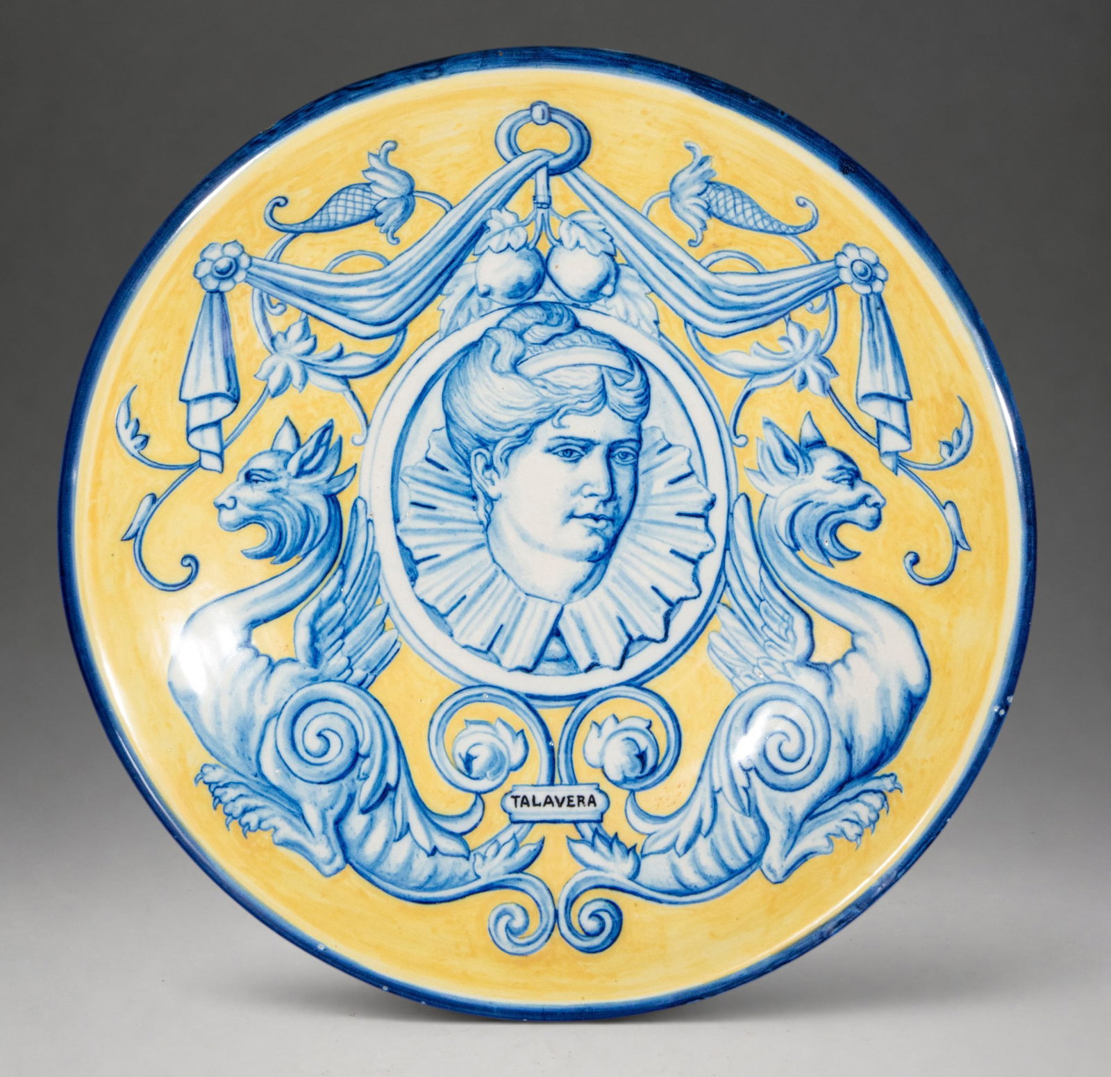 Talavera Niveiro decorative plate with female bust and taps – early 20th century: Circular ceramic plate, glazed and painted in blue and yellow. Diameter: 43 cm. Central decoration with a Renaissance-inspired female bust framed by a medallion, flanked by two facing griffins and acc