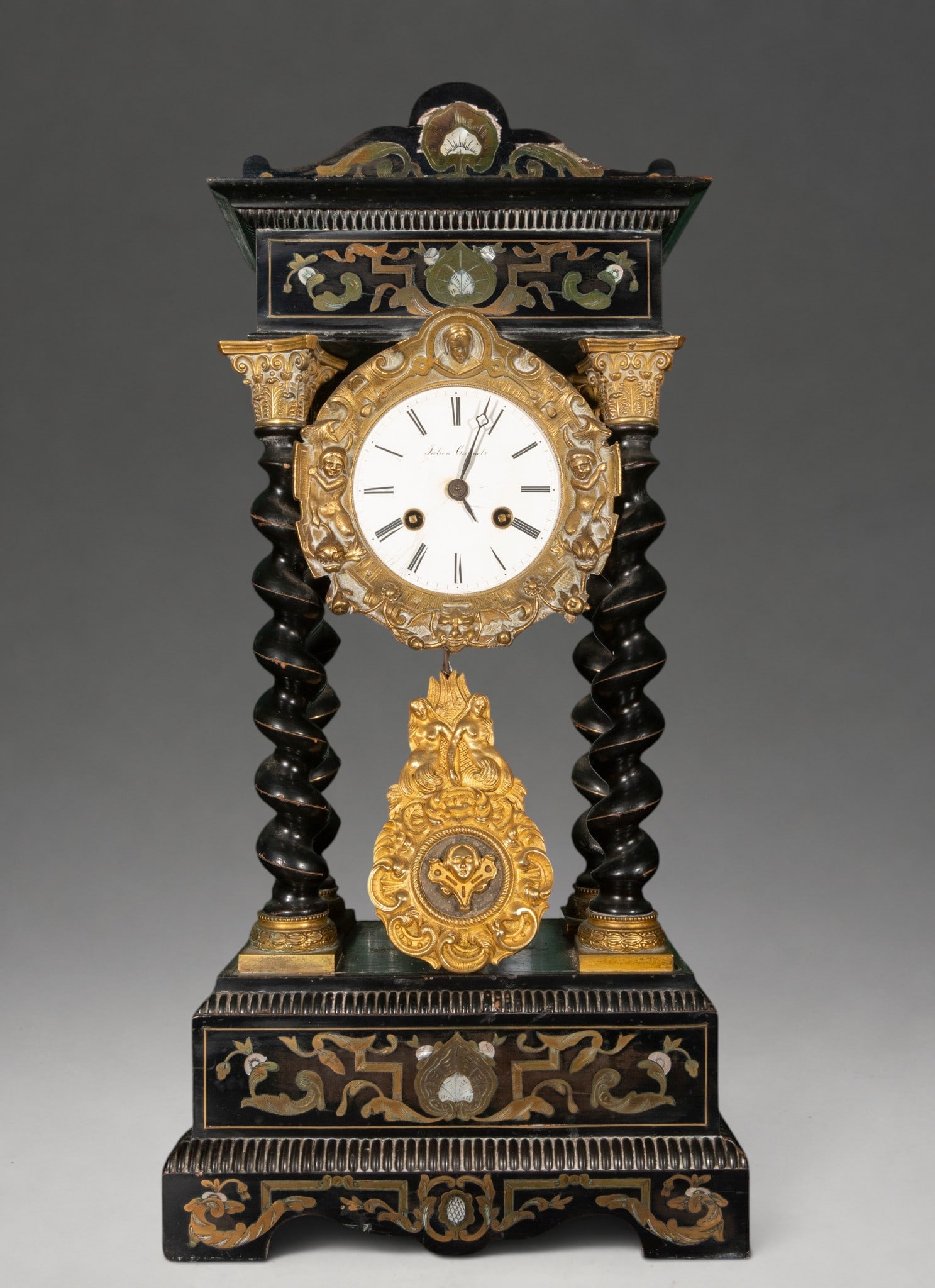 Napoleon III portico clock with Solomonic columns – 19th century: Ebonized wood portico clock with marquetry decoration and inlaid vegetal and floral motifs. Dimensions: 57 × 28 × 16 cm. Architectural structure supported by four Solomonic columns with gilt metal c