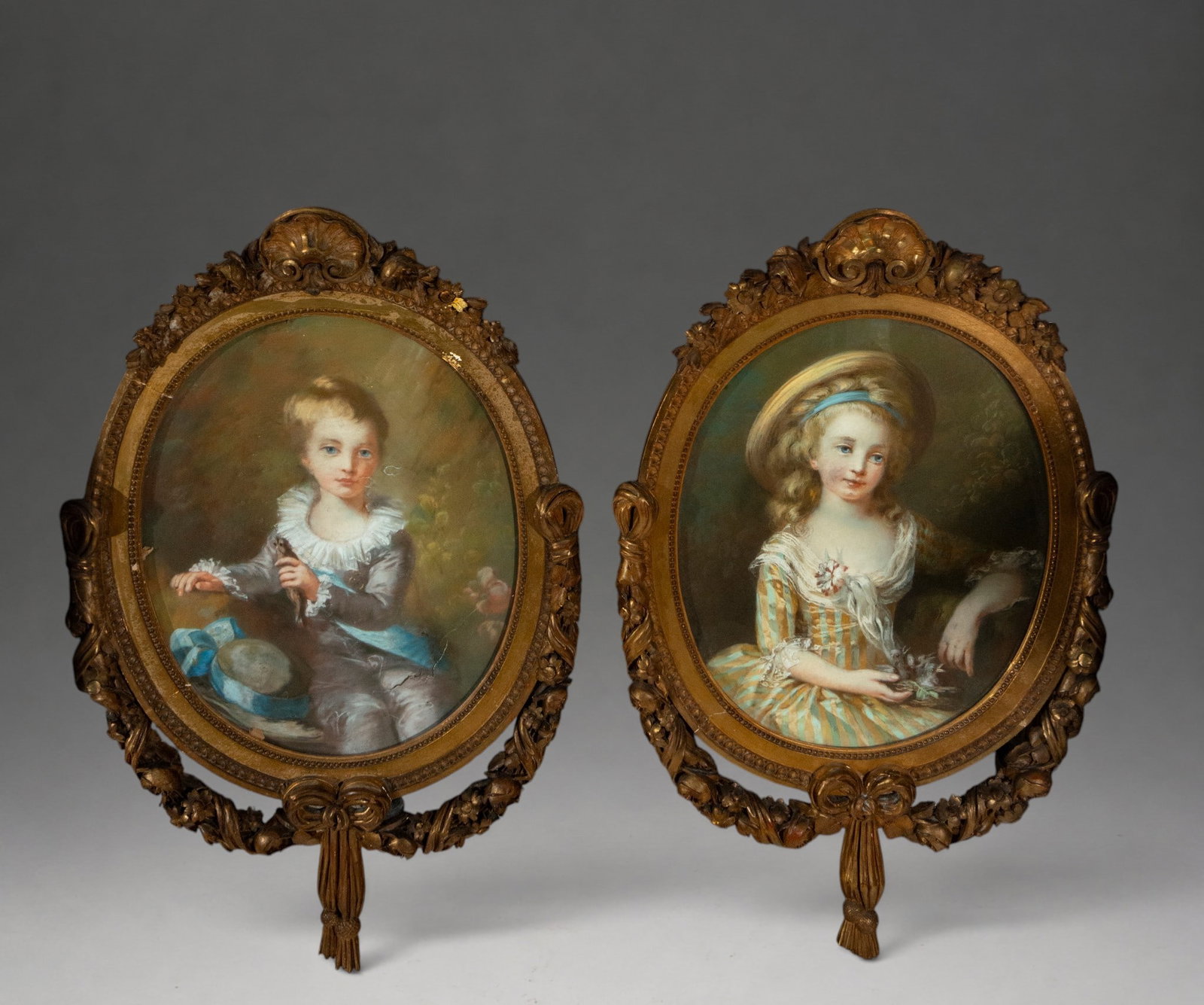 Pair of pastel portraits of children – European School – late 18th/19th century: A pair of pastel portraits of a young boy and girl, both half-length and set against landscape backgrounds. The boy wears a bluish-gray suit with a lace collar and blue sash, holding a small bird; bes