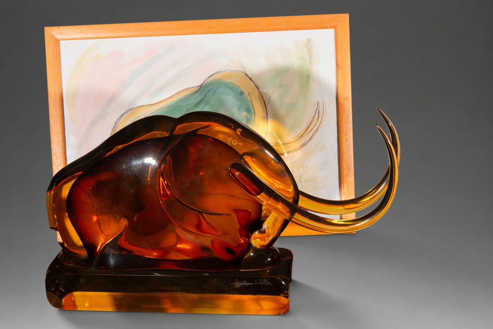 Mammoth in Murano glass, signed – Marco Polo Murano – 20th century: Murano glass sculpture in amber tones, depicting a reclining mammoth with voluminous forms and long, curved tusks. Signed on the base. Accompanied by a framed original sketch, signed and bearing the c