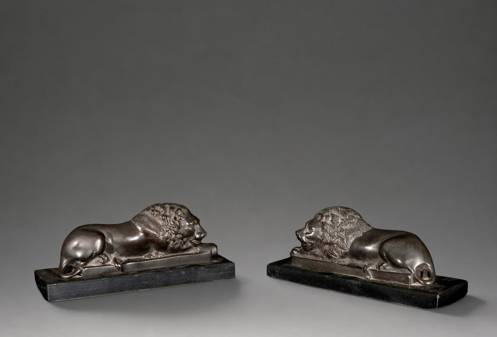 Pair of reclining lions – 19th century: Pair of patinated bronze sculptures depicting reclining and facing lions, modeled with abundant detail in their manes and mounted on rectangular black marble bases. This style is linked to the Grand T