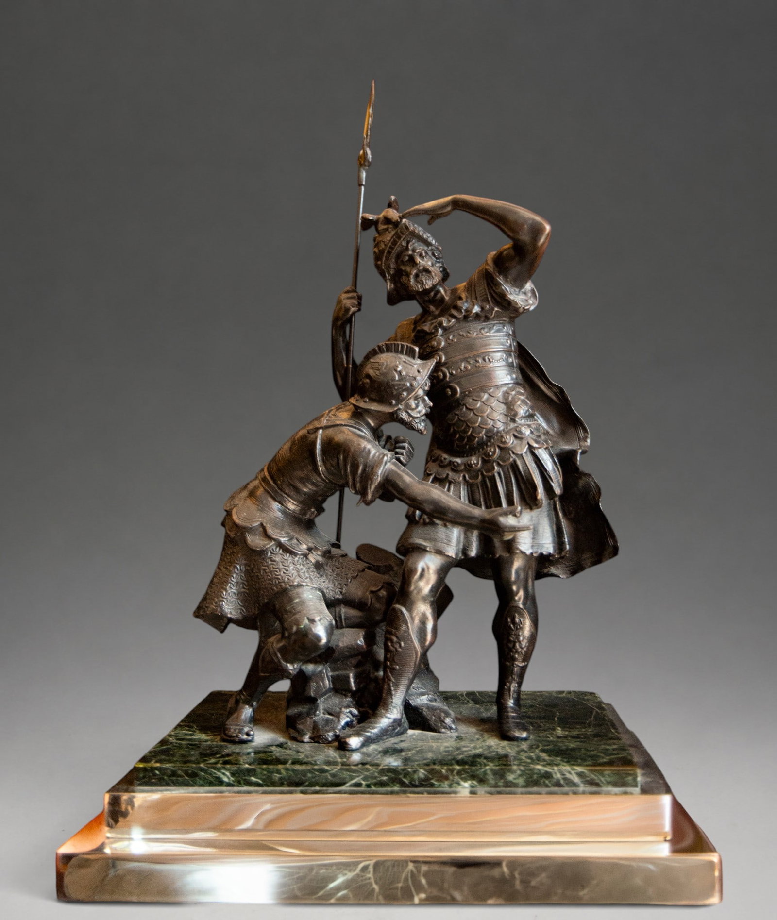 Pair of Warriors in bronze and marble - 20th Century European School: A patinated bronze sculpture group depicting two warriors in a historical scene. The main figure, standing and clad in armor, helmet, and cloak, holds a long spear while raising his left hand to his f