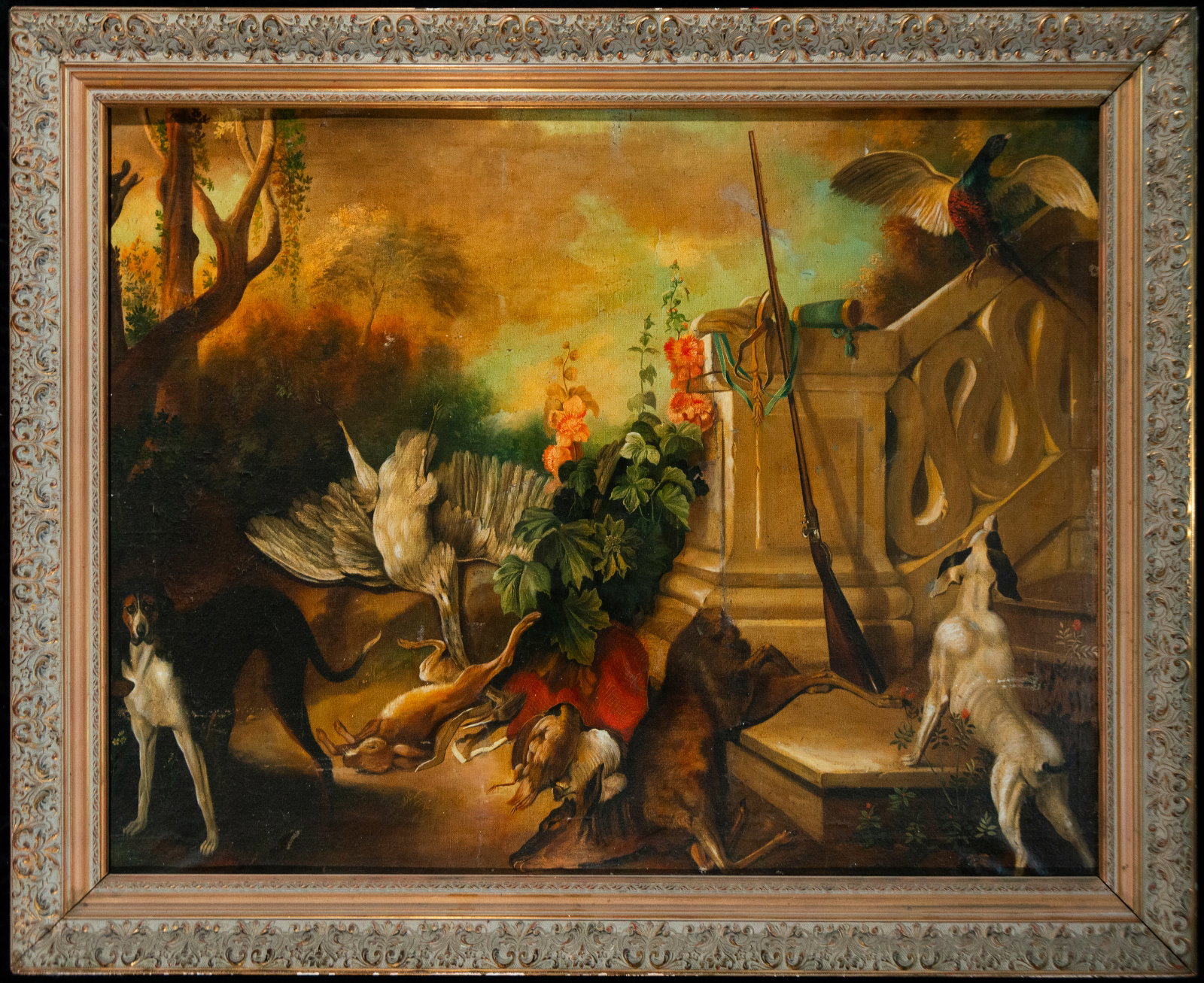 Hunting still life with dogs and birds – Spanish School – 20th century: Oil on canvas. Hunting scene with two dogs, small game, and birds, including a pheasant, accompanied by a shotgun, game bag, and other hunting equipment, set against a wooded landscape. A decorative c